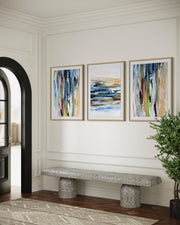 Three abstract paintings on a wall above a stone bench in a room with a plant and open door.