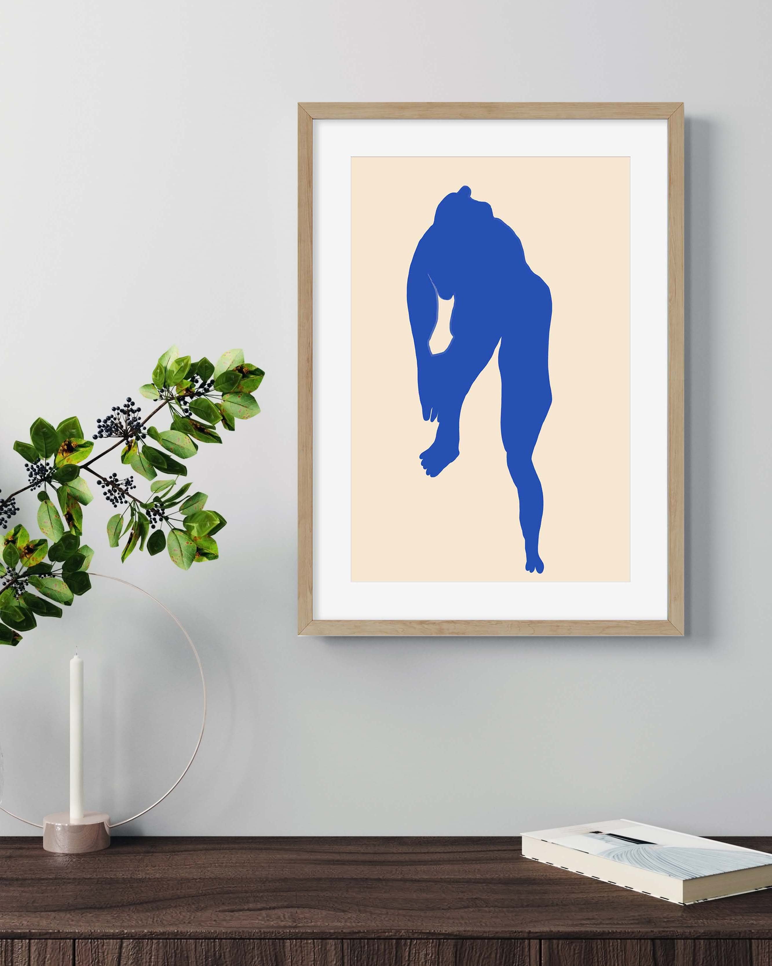 Framed artwork of a blue silhouette on a beige background, displayed on a wall above a wooden surface with a plant and books.