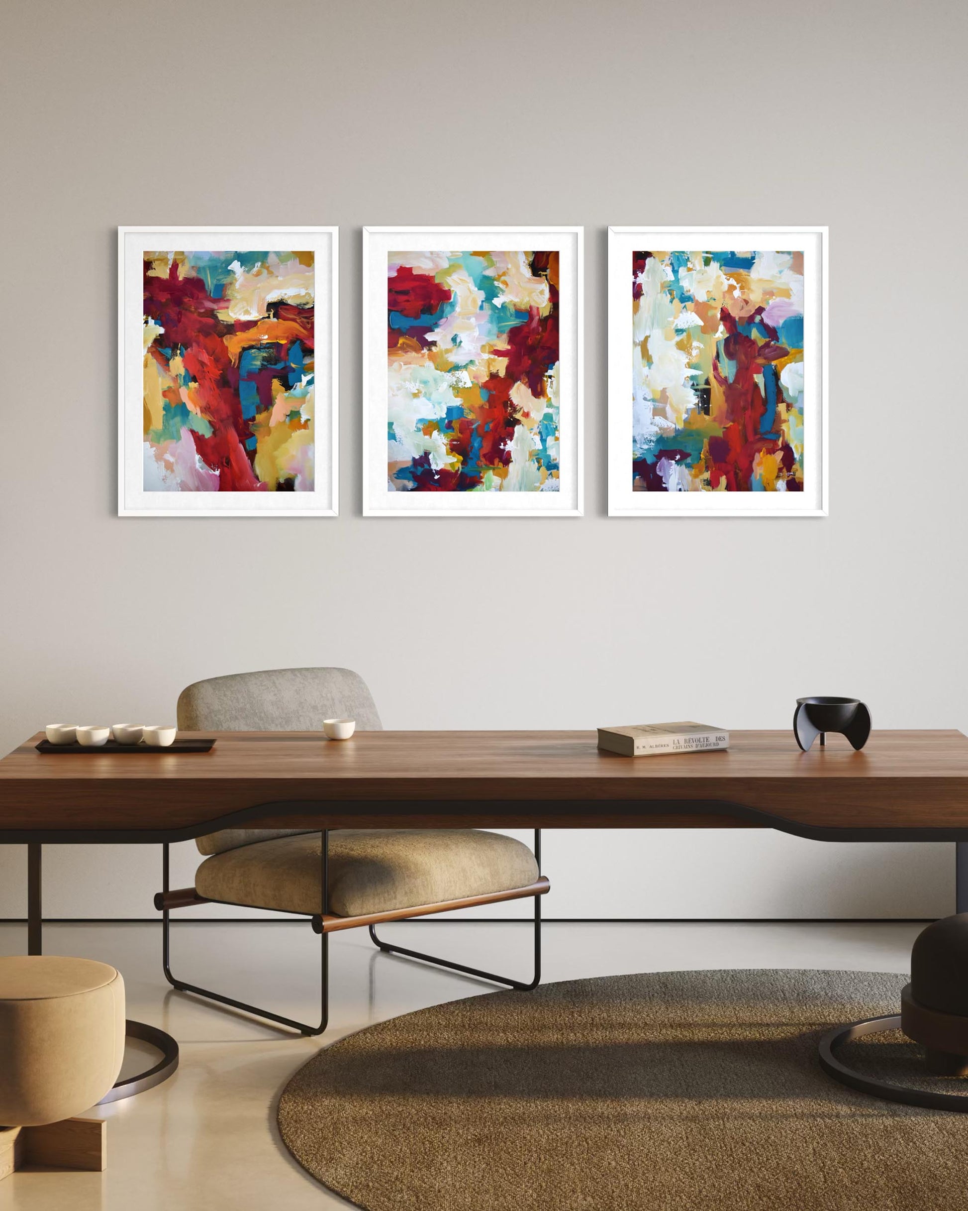 Three abstract paintings in white frames on a wall above a wooden table.