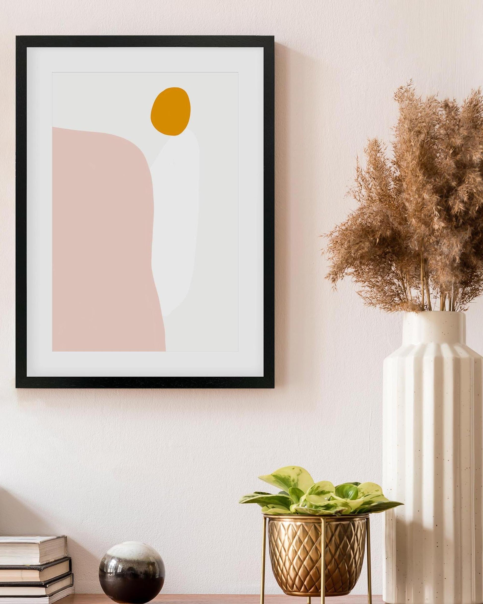 Framed abstract art with pink and yellow shapes on a wall next to a vase with dried flowers.