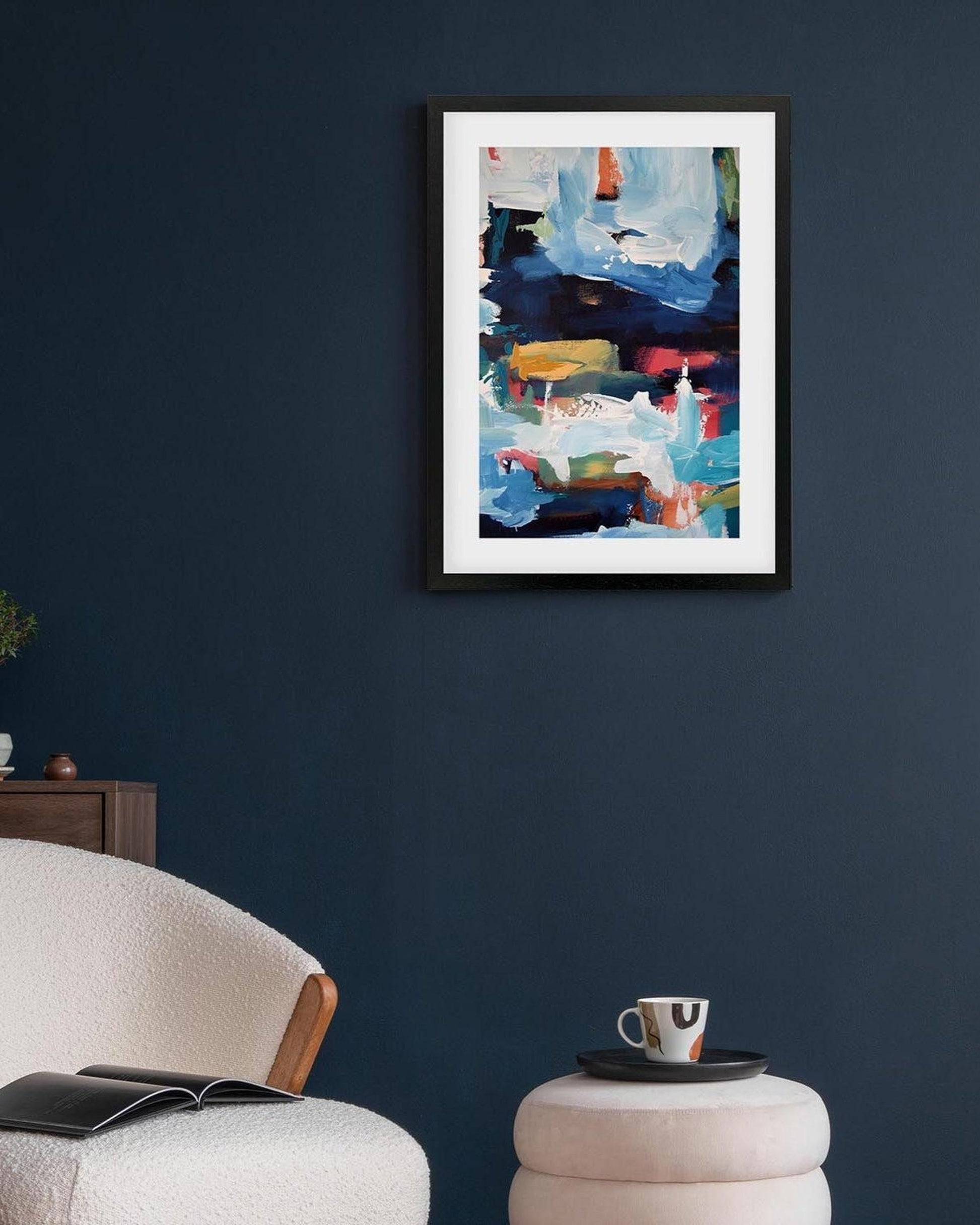 Abstract painting on a dark blue wall above a white armchair with a book and a small table with a cup.