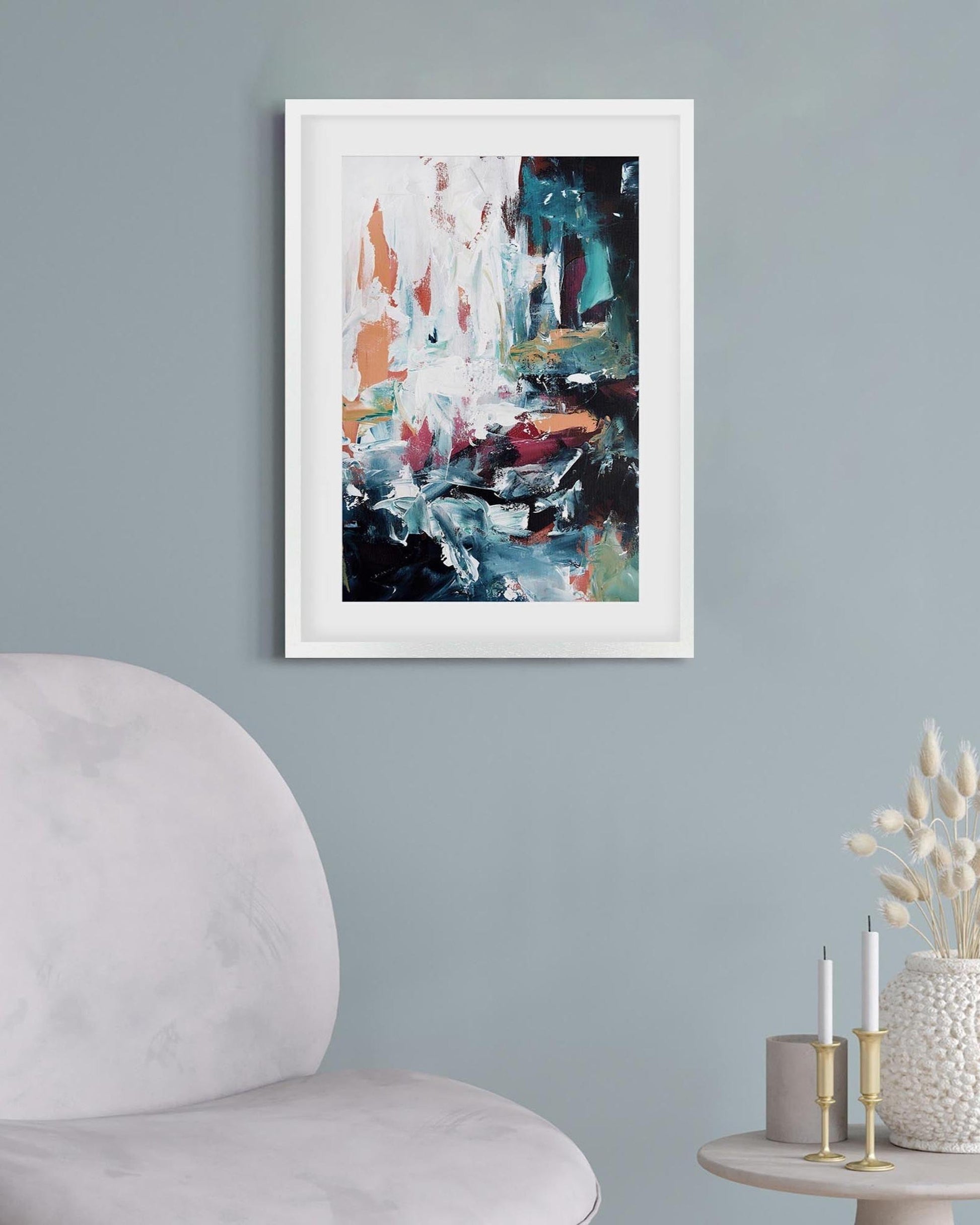 Abstract painting in a white frame on a gray wall above a chair and side table with decorative items.