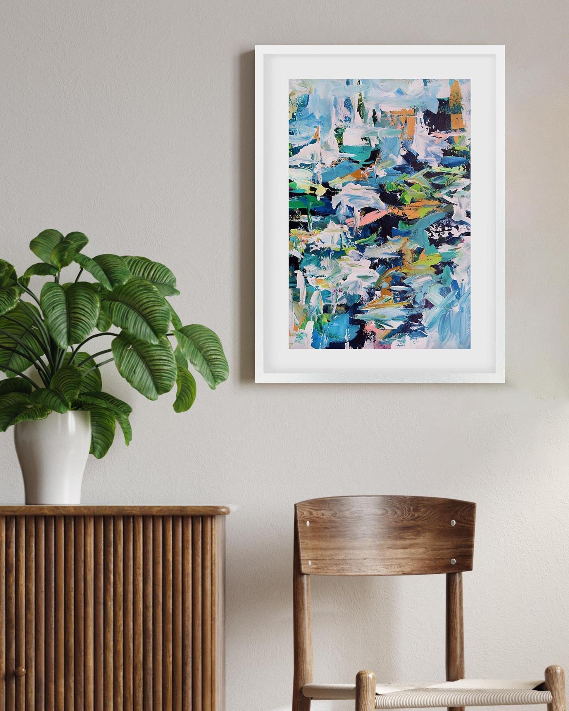 Framed abstract art piece on a wall with a plant and wooden furniture in the foreground.