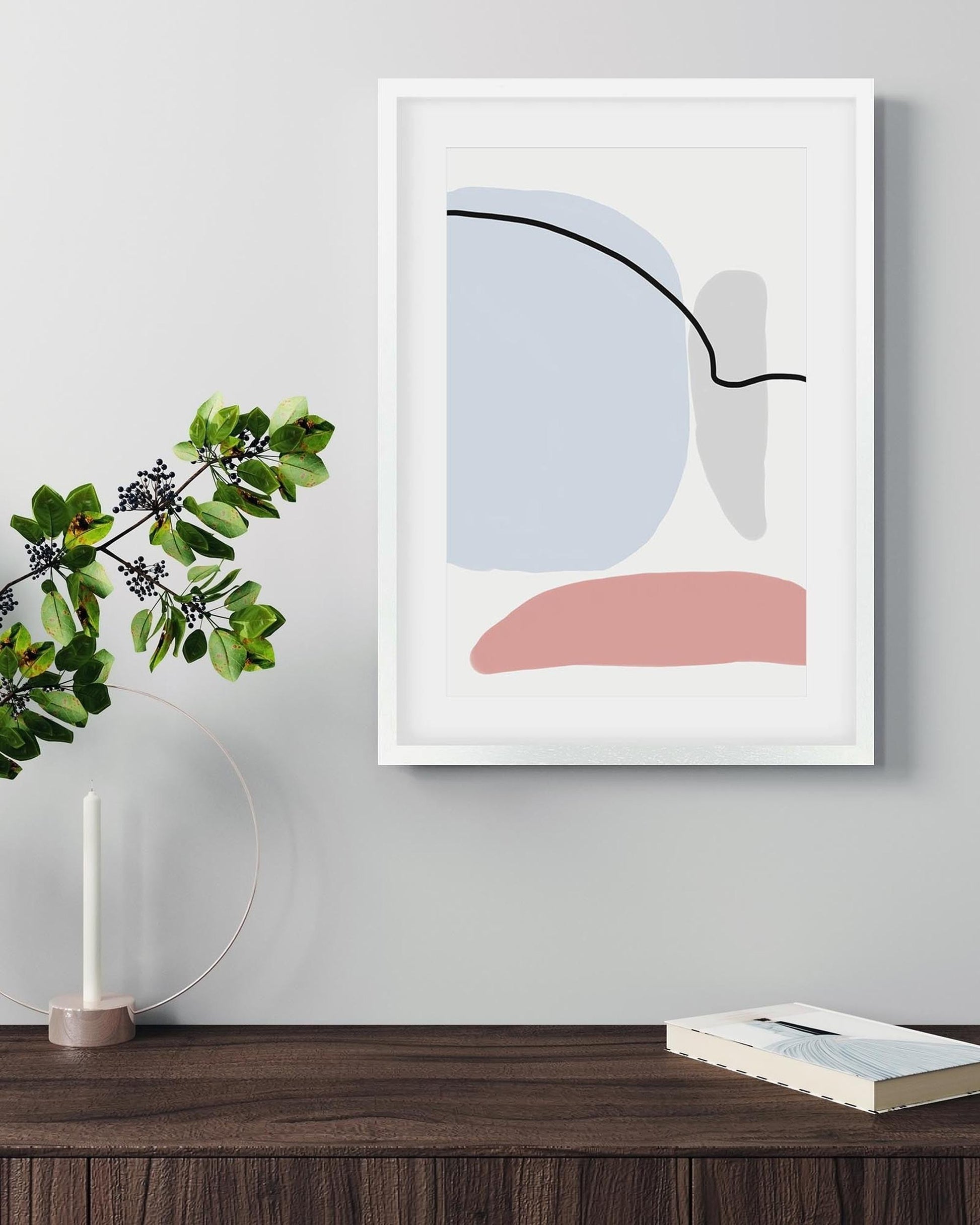 Abstract art print in a white frame on a wall above a wooden surface with a plant and book.