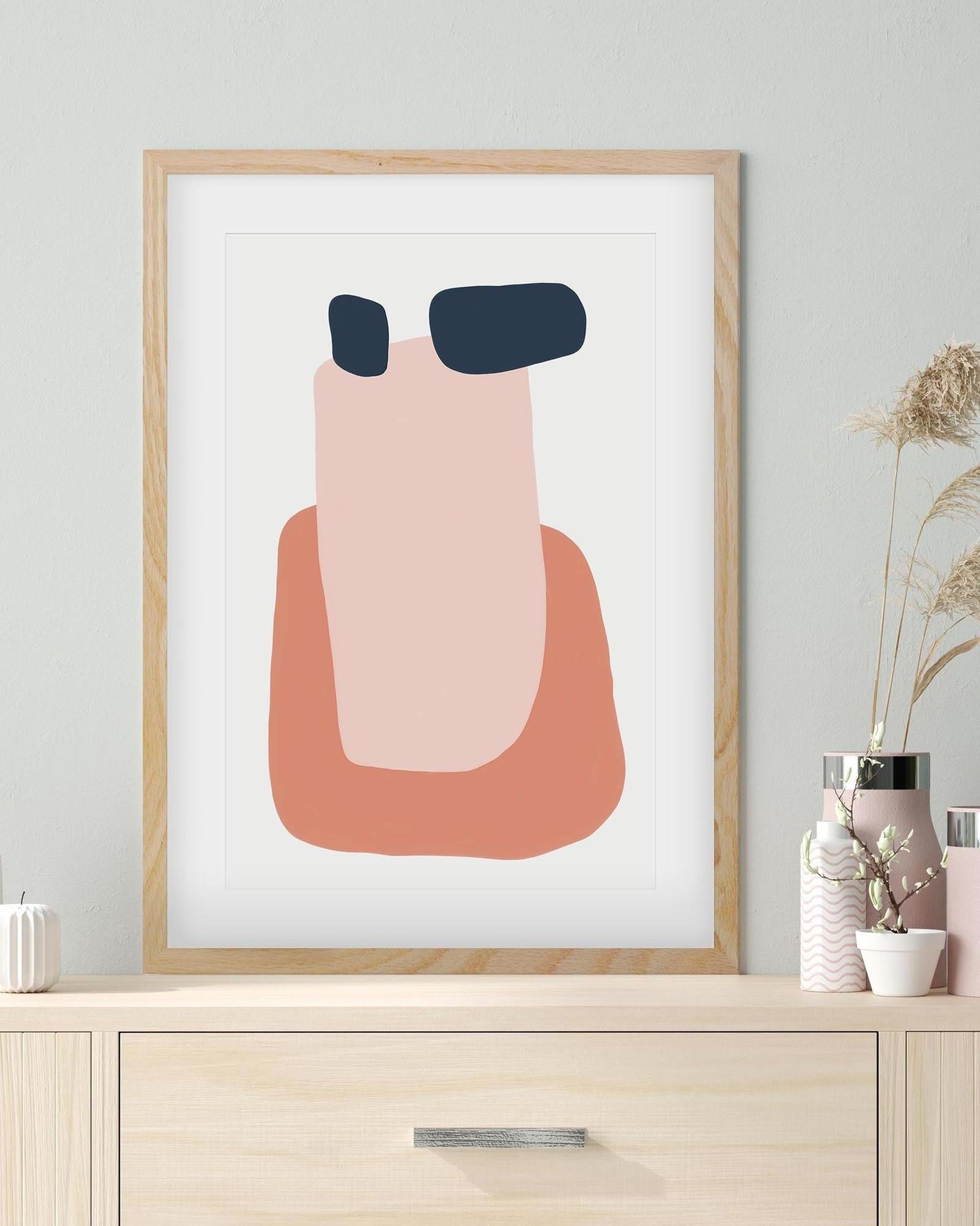 Framed abstract art print with pink and blue shapes on a white background, displayed on a wooden shelf.