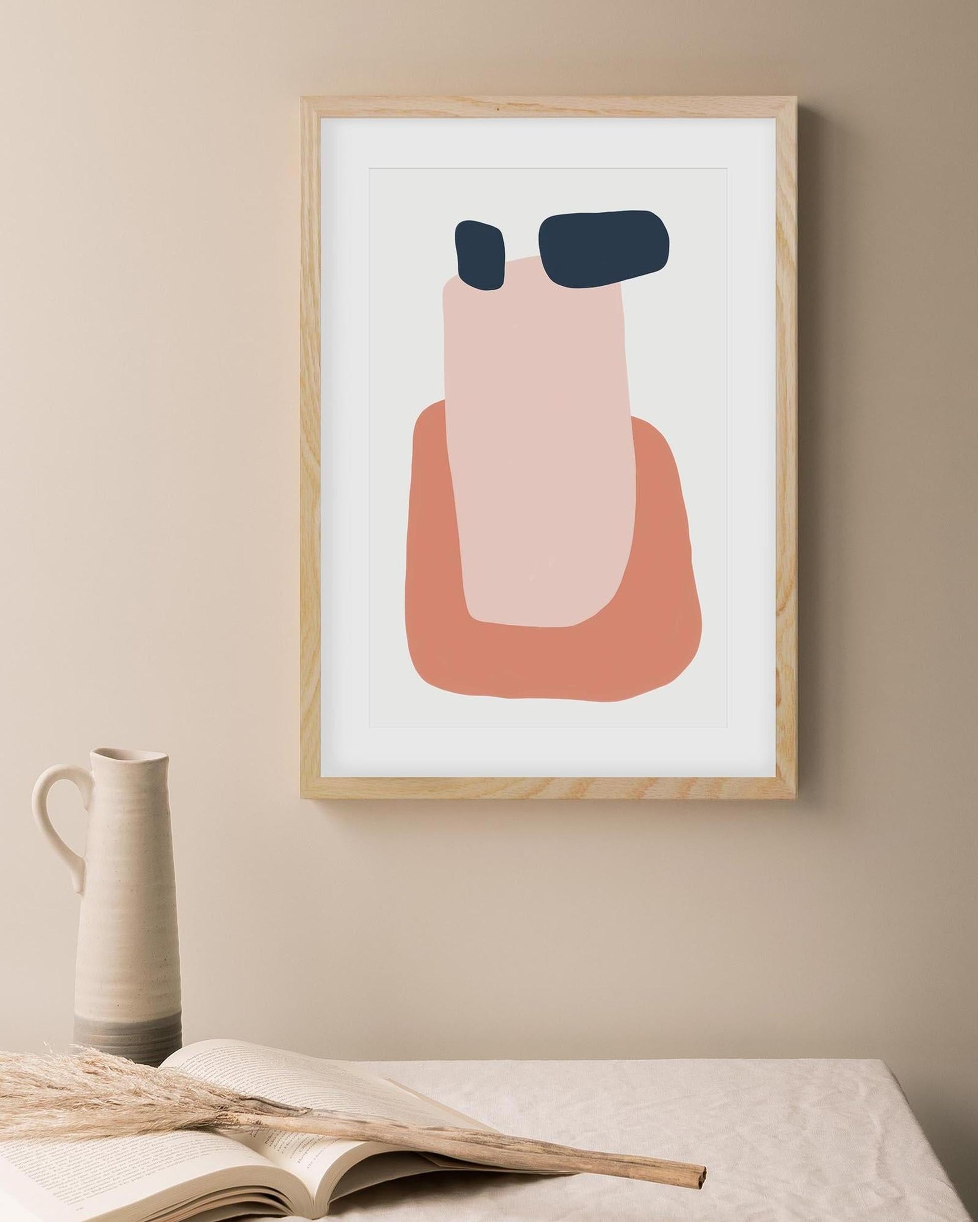 Abstract art print in a wooden frame on a beige wall with a vase and book below.