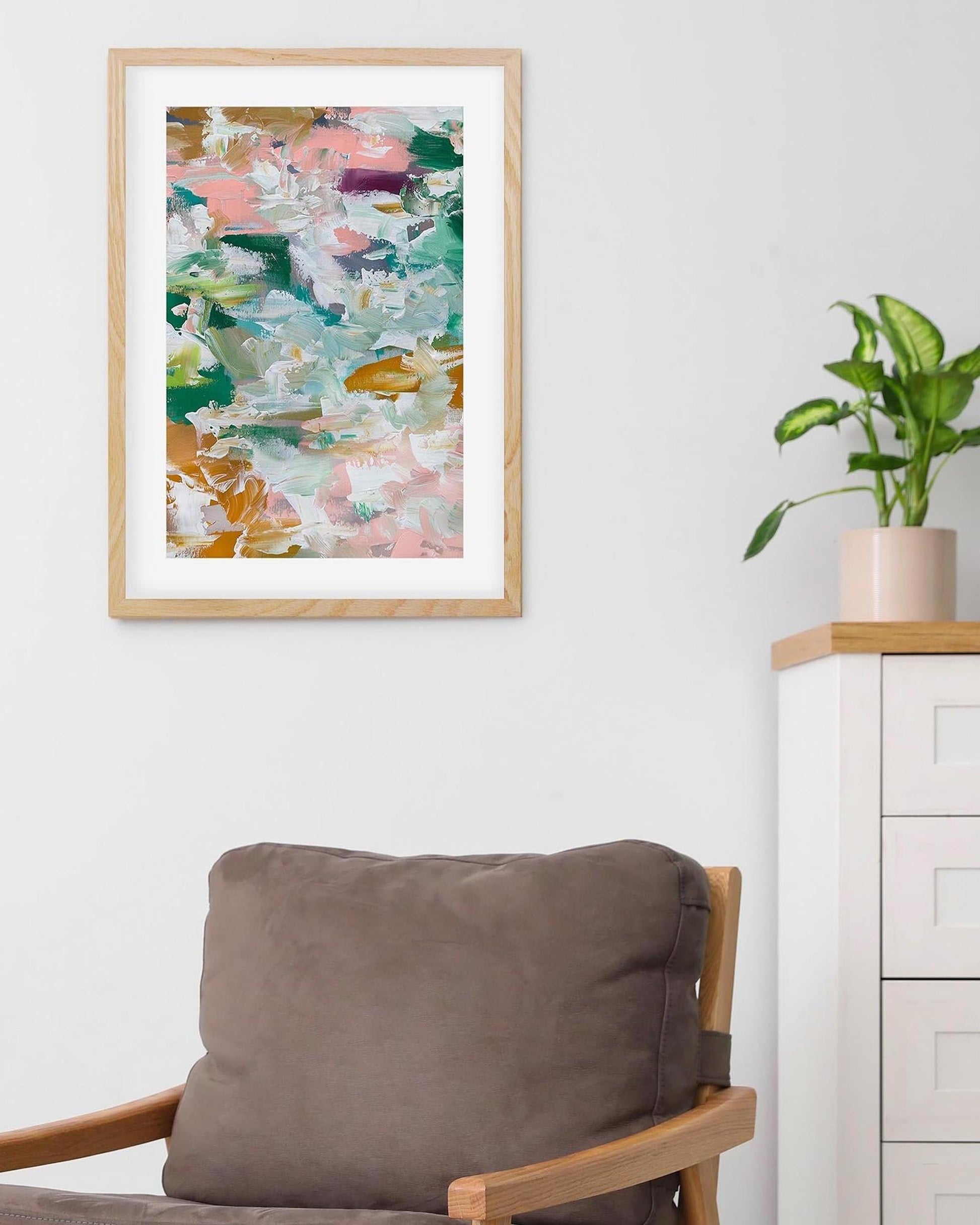 Framed abstract art piece on a wall above a chair with a plant in the background.