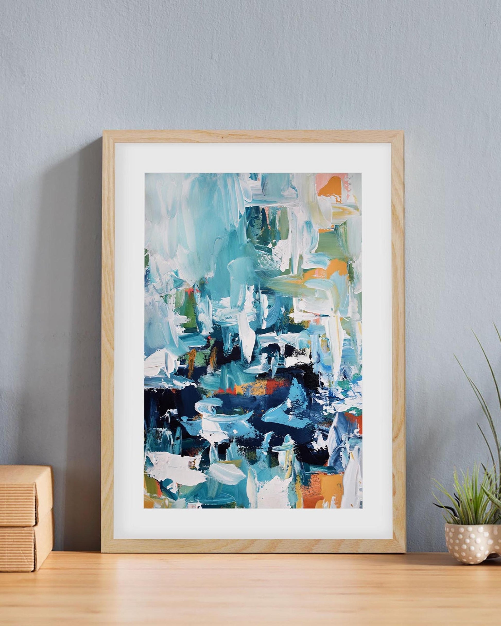 Framed abstract art print on a wall with a plant and box in the background