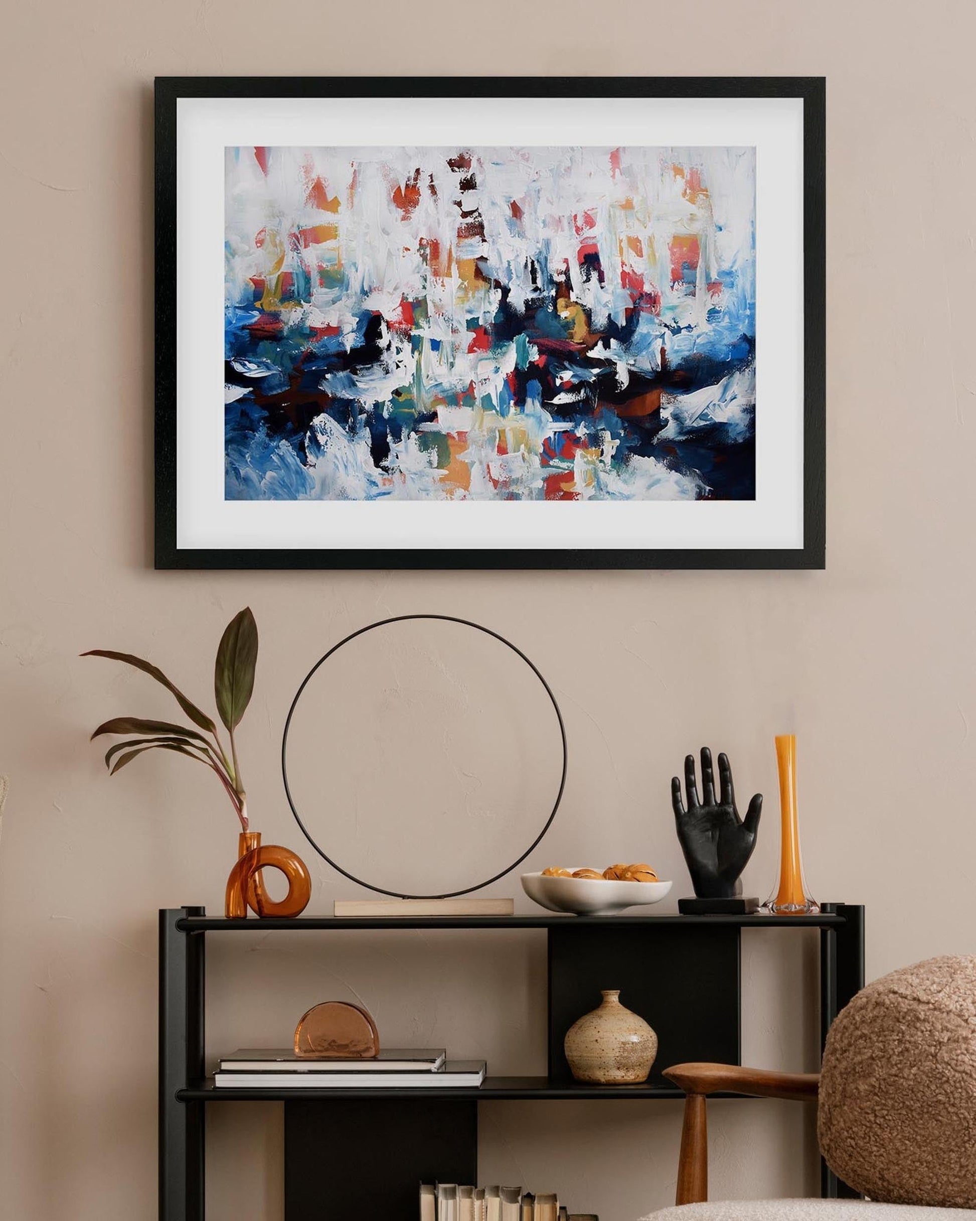 Abstract painting in a black frame on a beige wall above a shelf with decorative items.