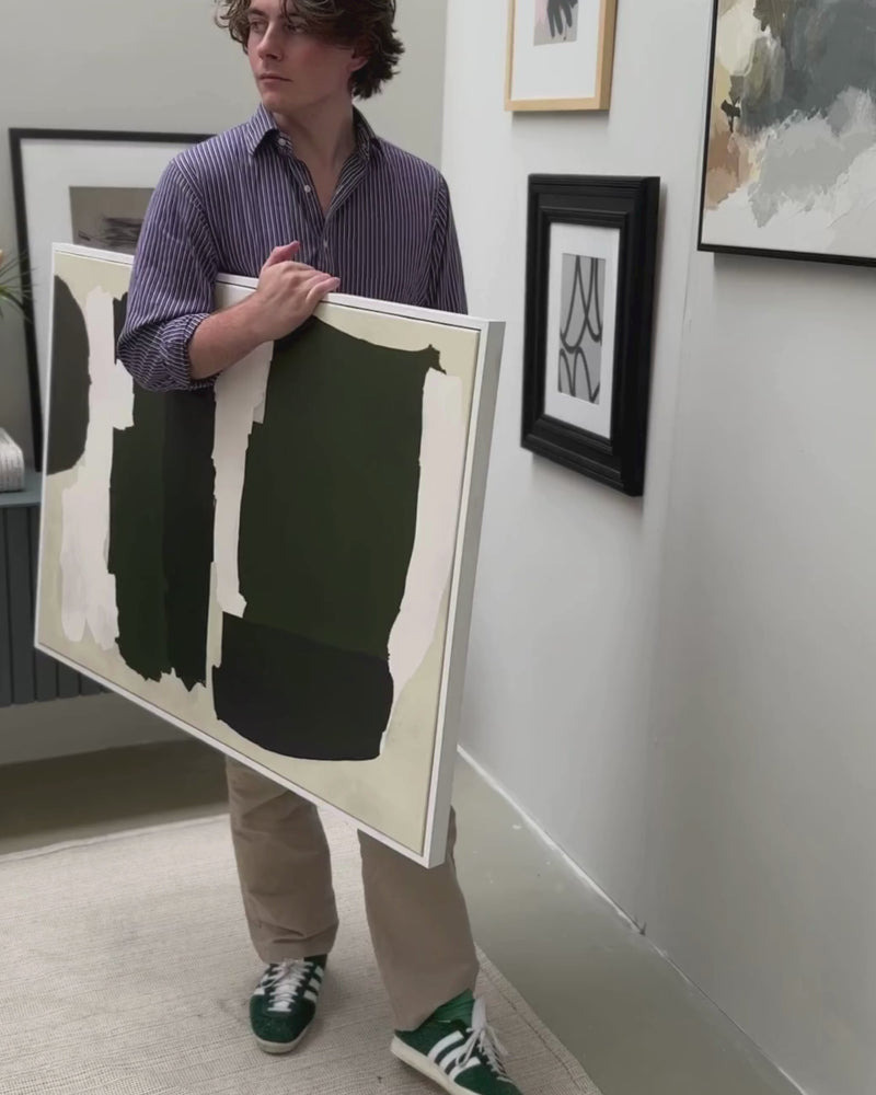 Video of art lover holding a large framed canvas with abstract shapes in green and neutral palette ready to hang the piece on a wall.