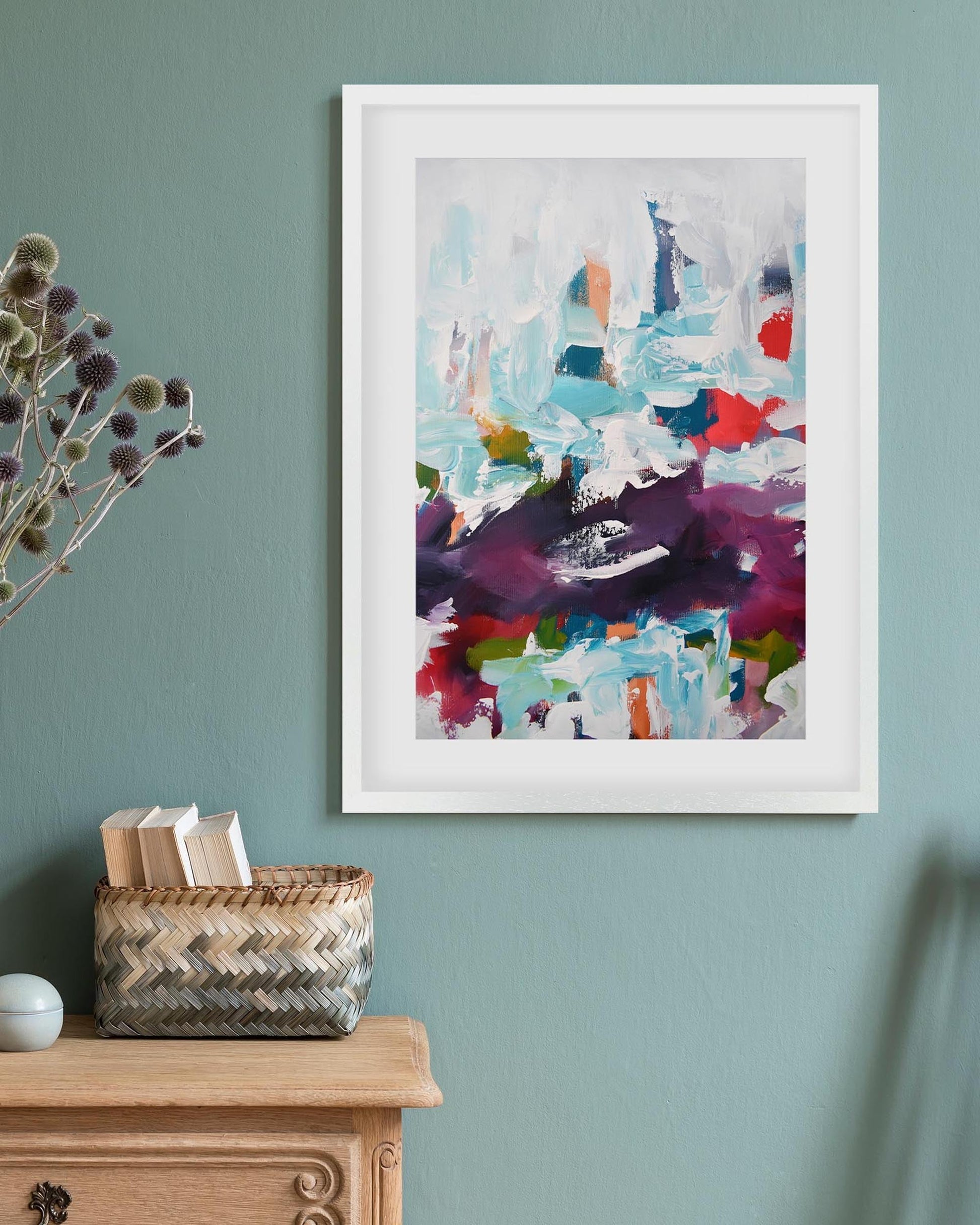 Framed abstract art on a wall with a vase and basket on a wooden surface below.