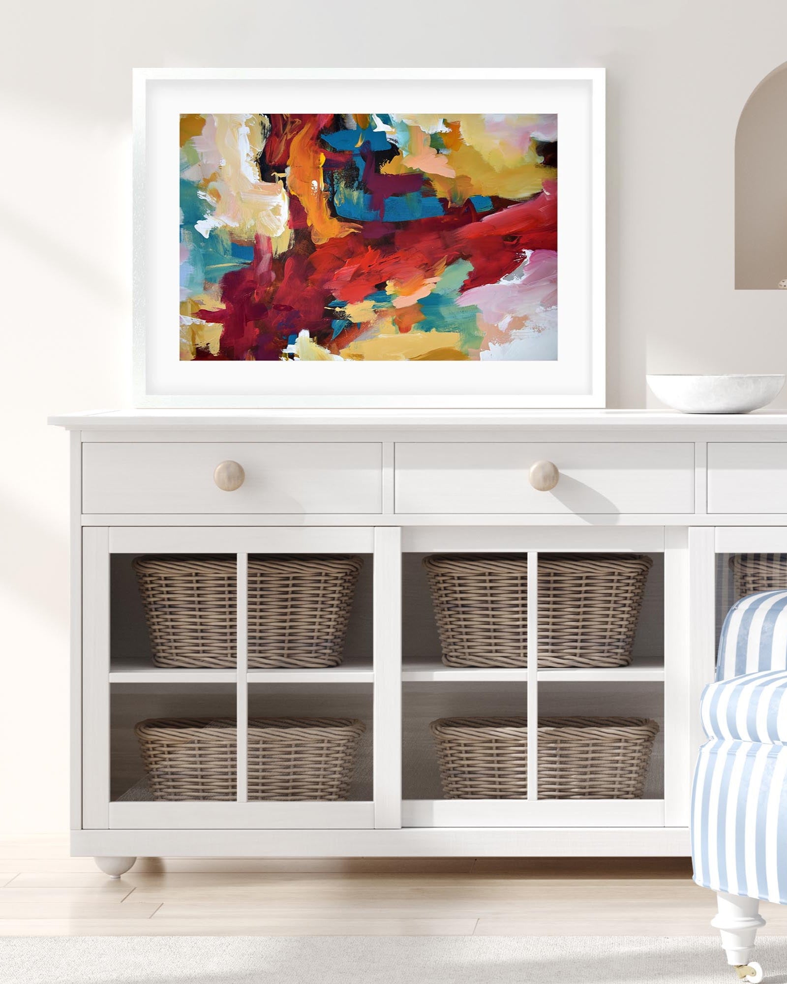 Colorful abstract painting on a white wall above a white cabinet with wicker baskets.