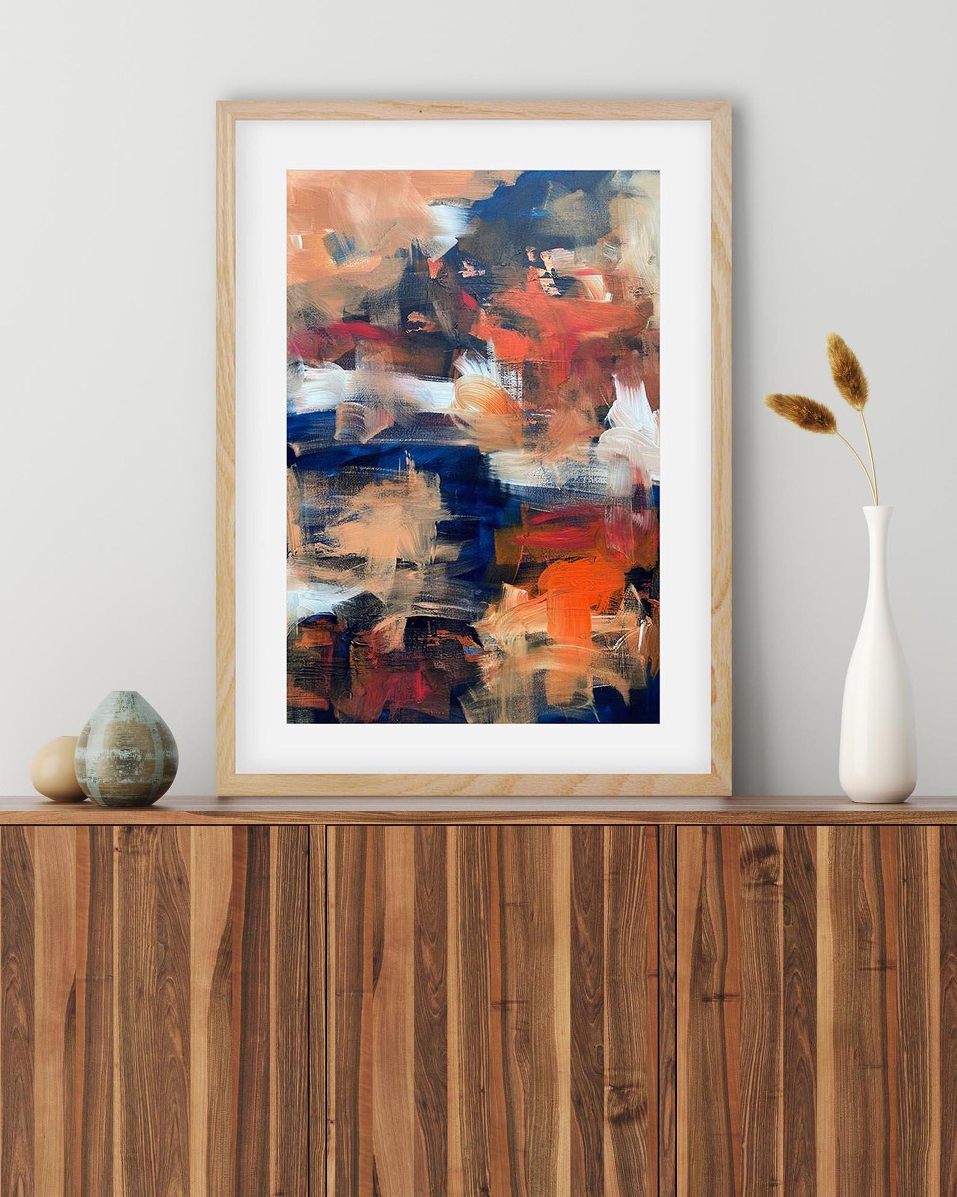 Abstract art print in a wooden frame on a wall above a wooden shelf.
