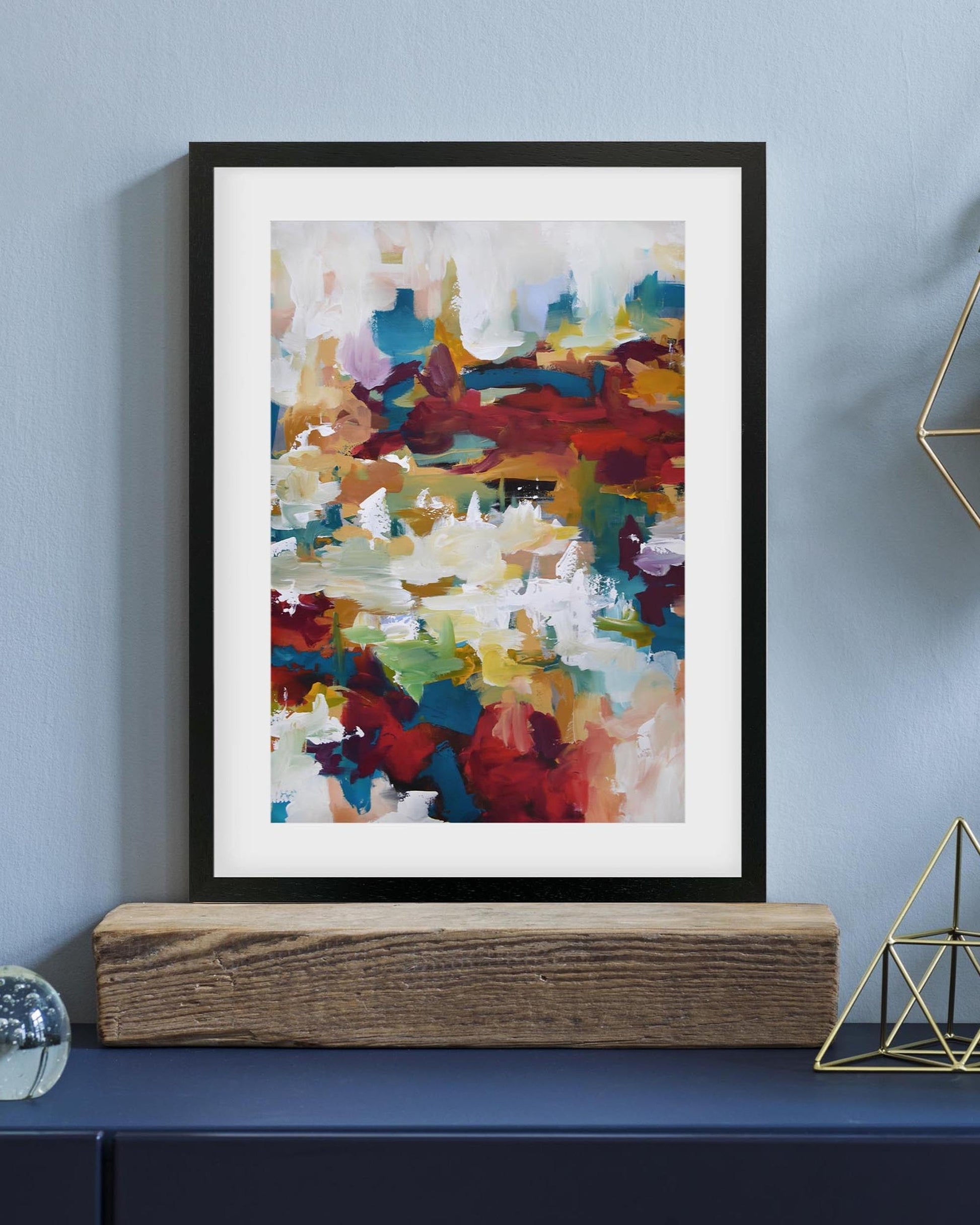 Framed abstract art print on a wall above a wooden shelf.