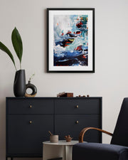 Framed abstract painting above a black dresser in a room with a plant and chair.