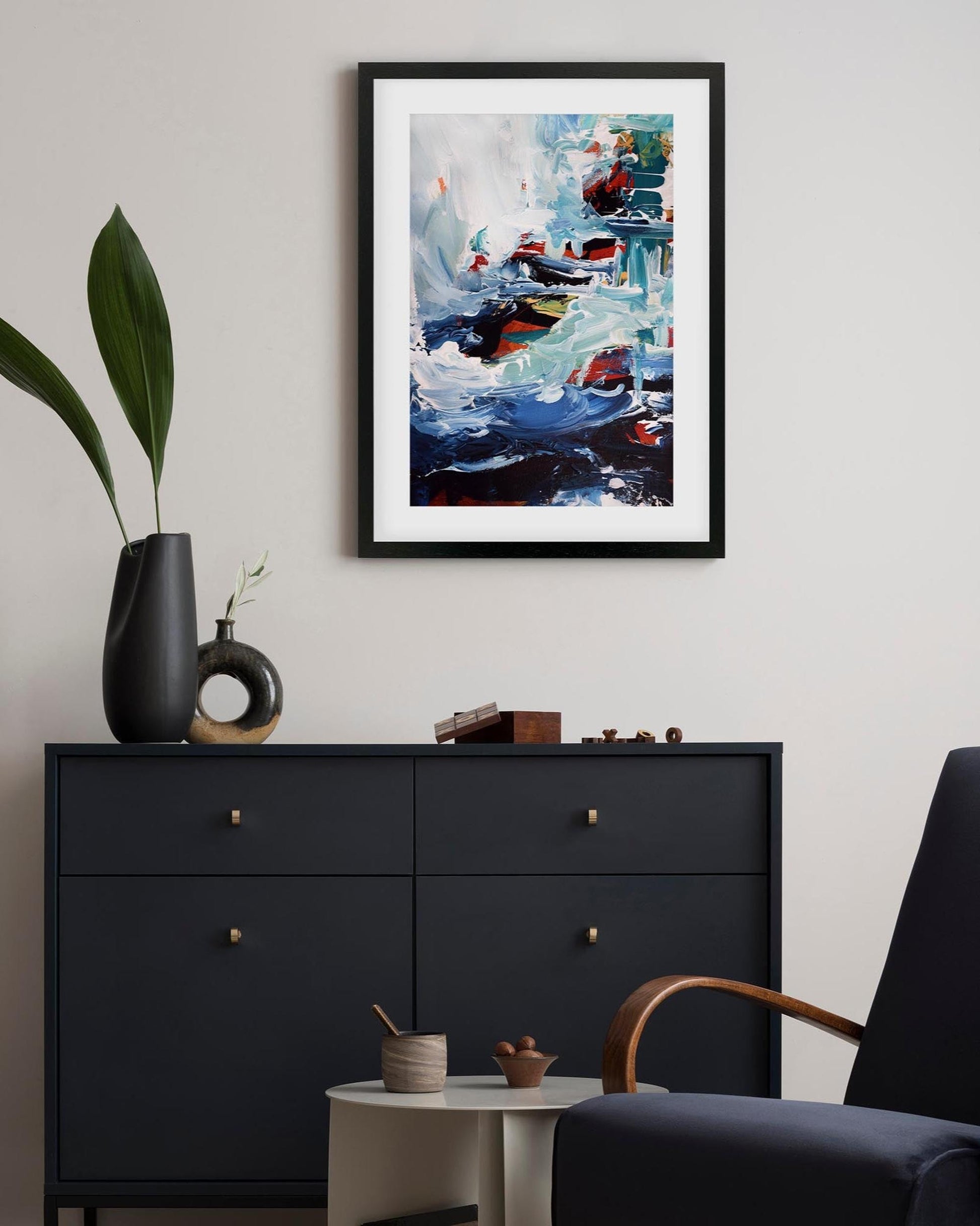 Framed abstract painting above a black dresser in a room with a plant and chair.