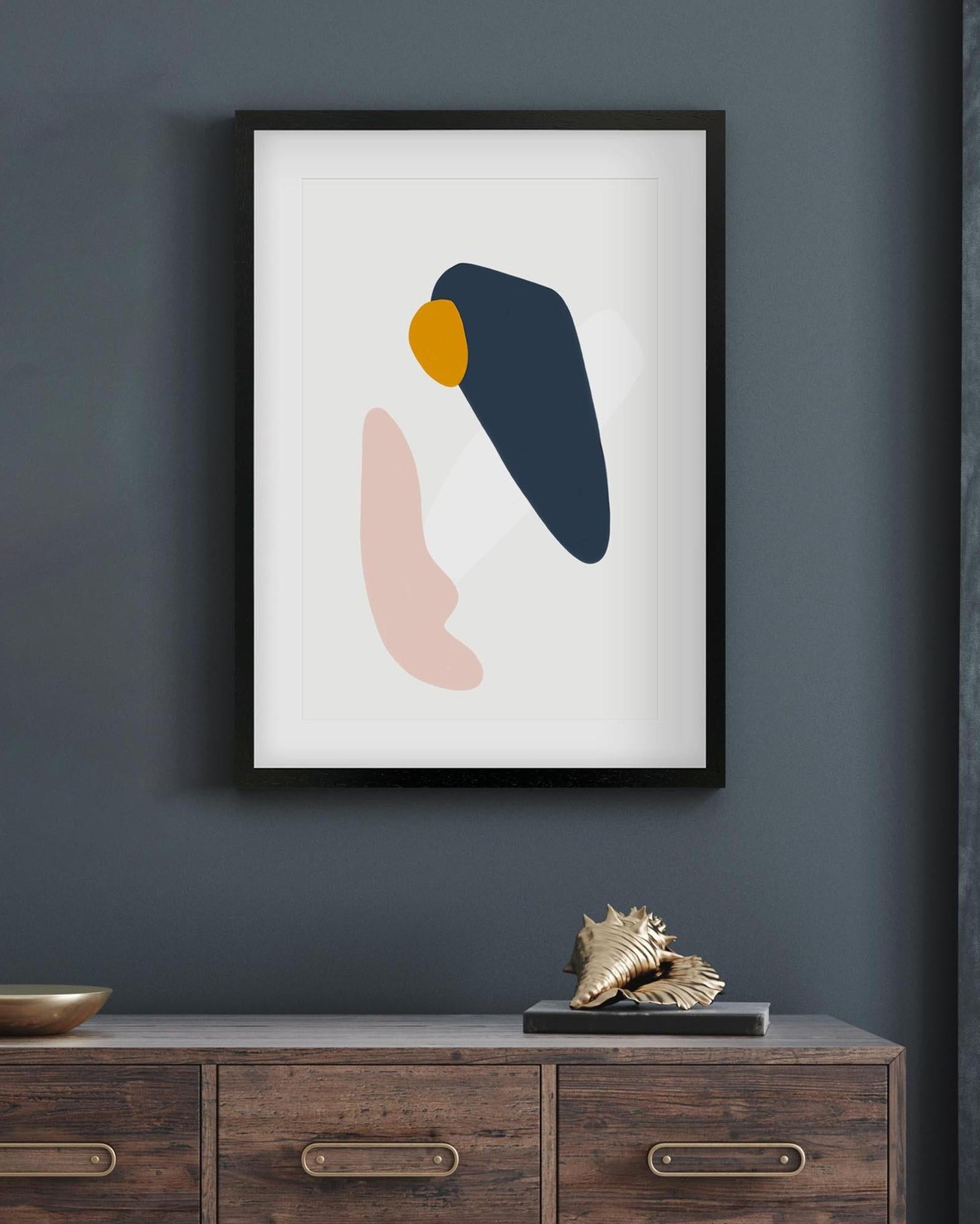 Framed abstract art print with blue, pink, and yellow shapes on a dark gray wall above a wooden cabinet.