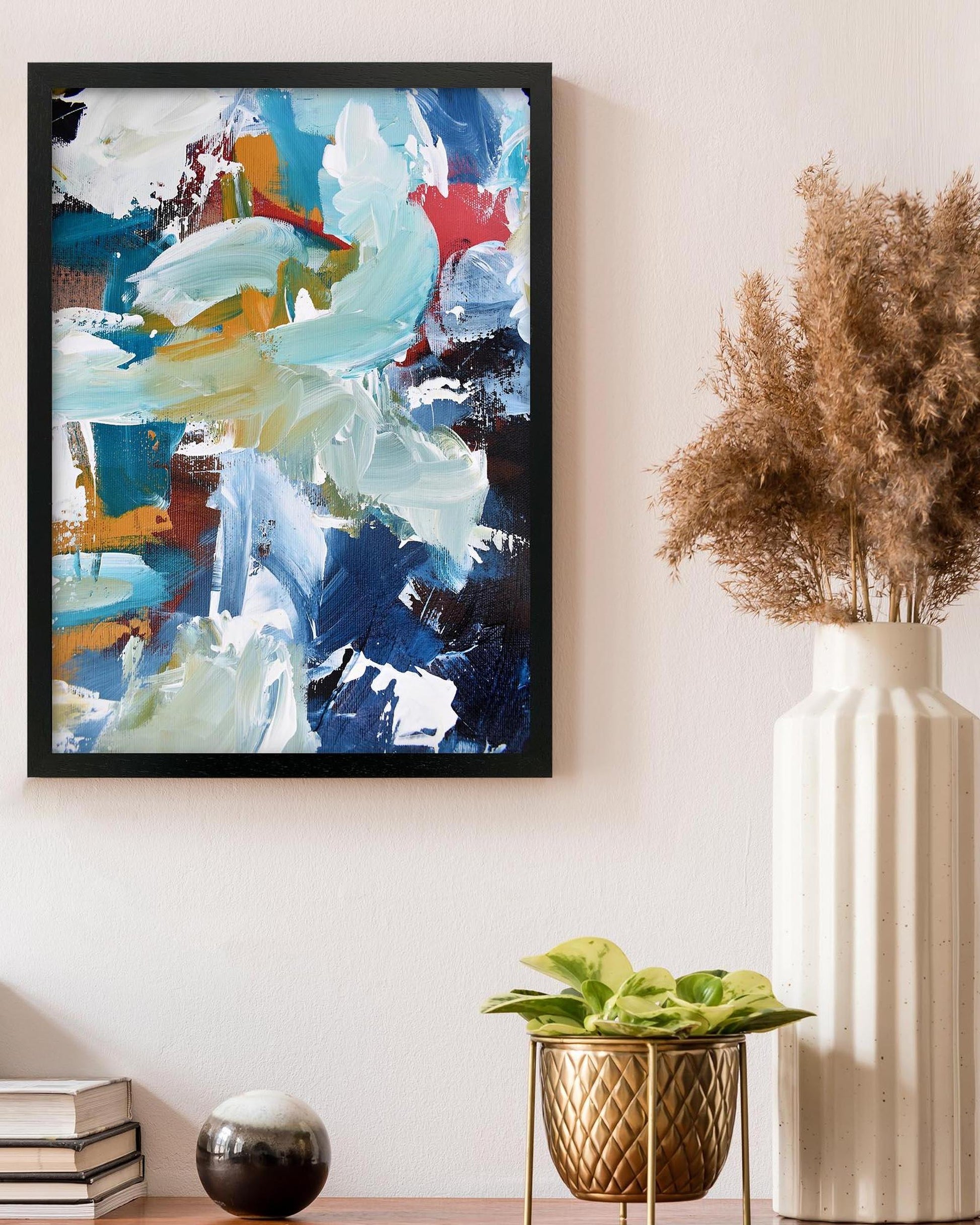 Abstract painting in a black frame on a wall with decorative plants and books below.