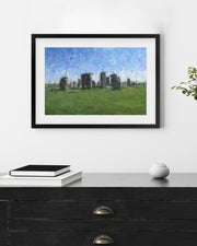Framed artwork of stone structures in a field on a white wall above a wooden surface.