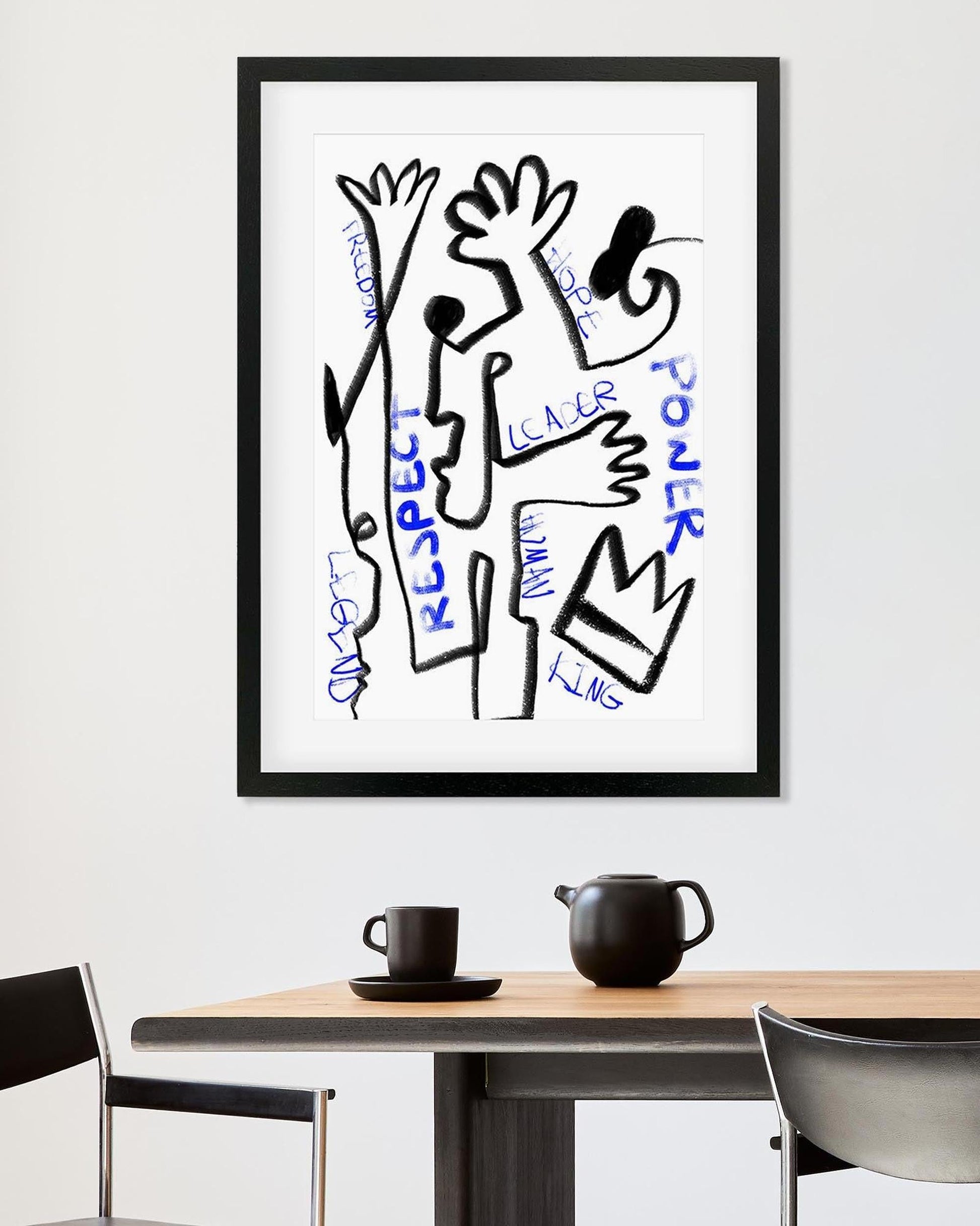 Framed abstract artwork with text on a wall above a table with a cup and teapot.