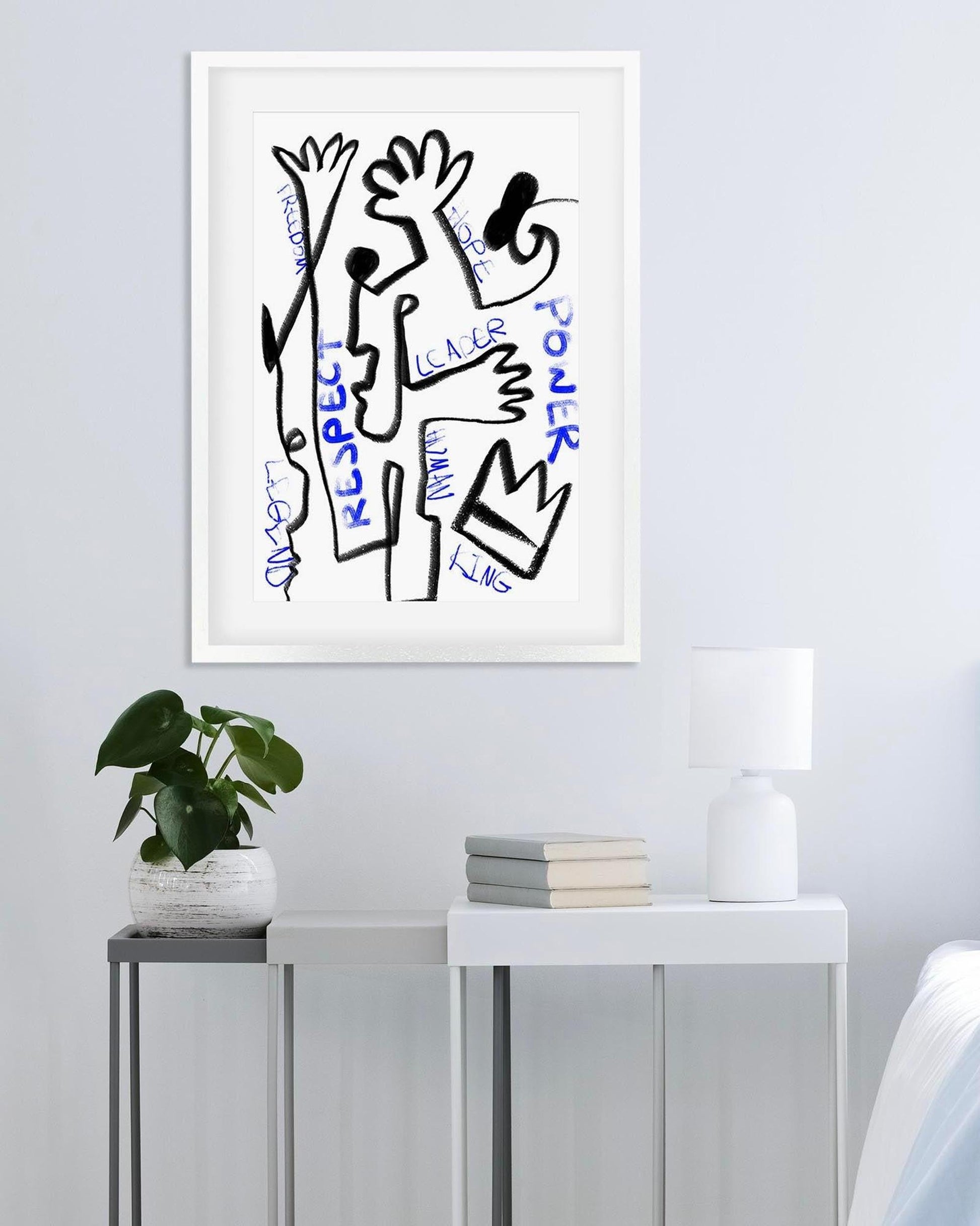 Framed abstract artwork with text on a wall above a small table with books and a plant.