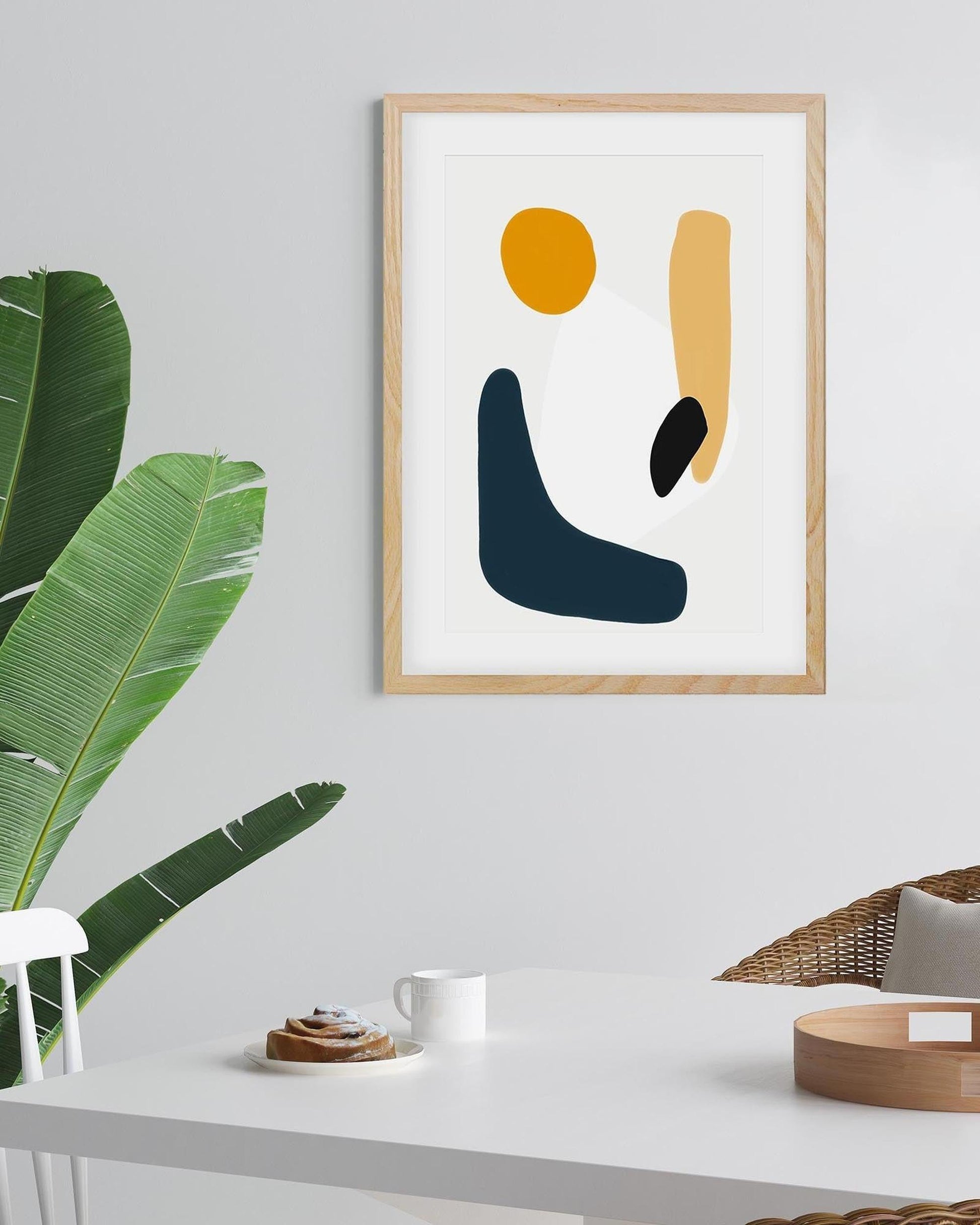 Abstract art print in a wooden frame on a wall with a plant and table in the foreground.