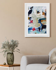 Framed abstract painting on a beige wall above a white armchair with a vase of flowers on a side table.