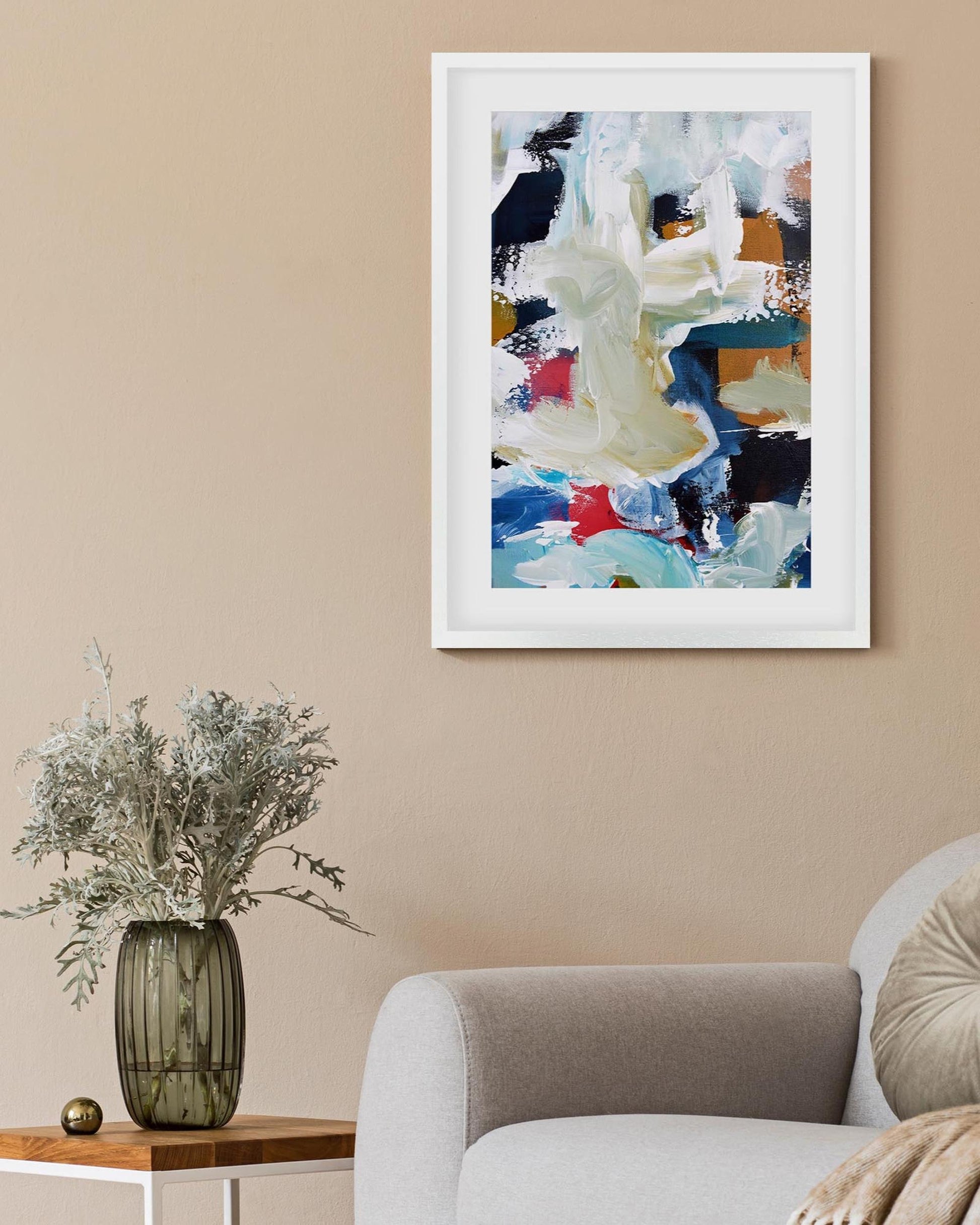 Framed abstract painting on a beige wall above a white armchair with a vase of flowers on a side table.