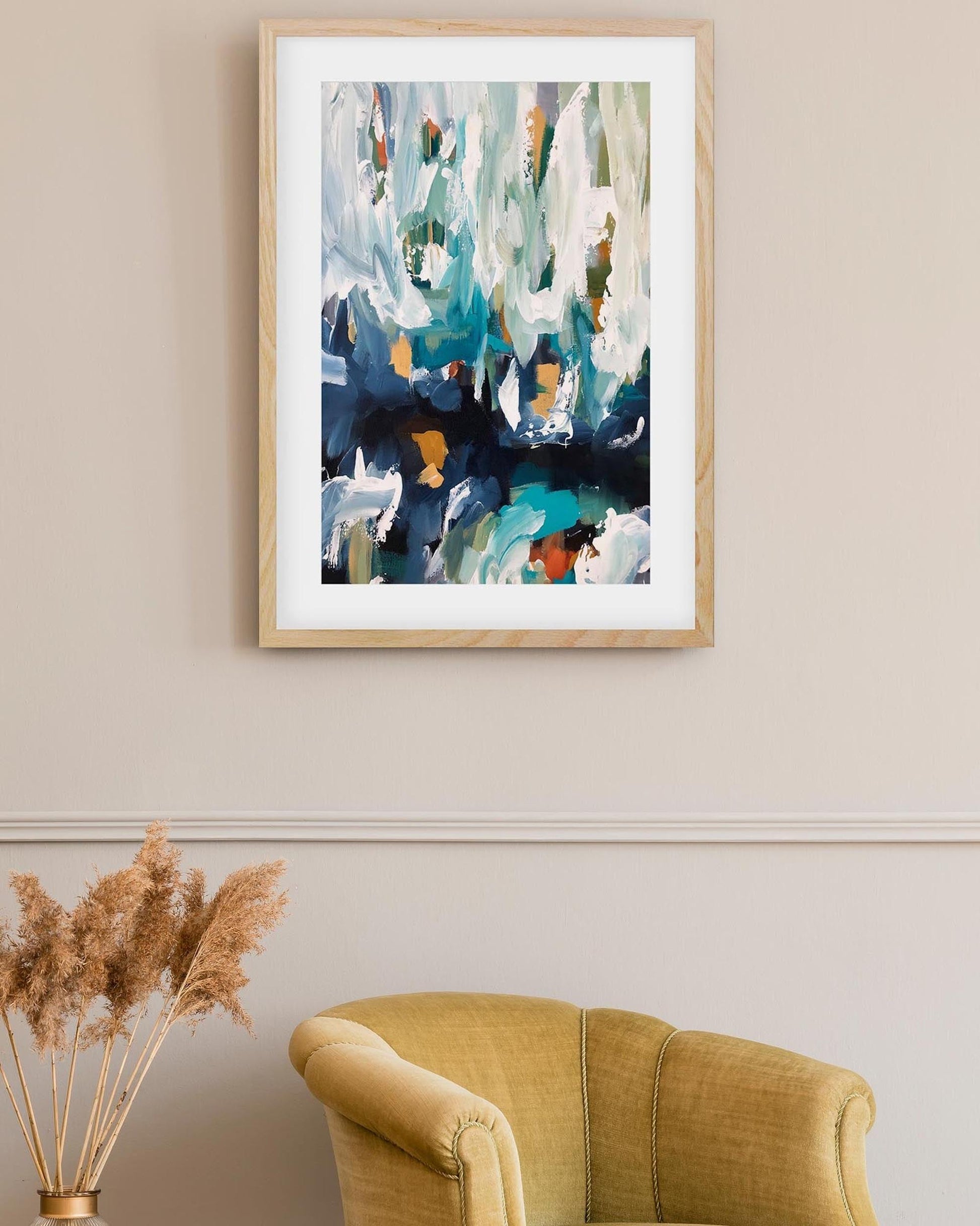 Framed abstract painting on a wall above a yellow armchair and dried plant decor.