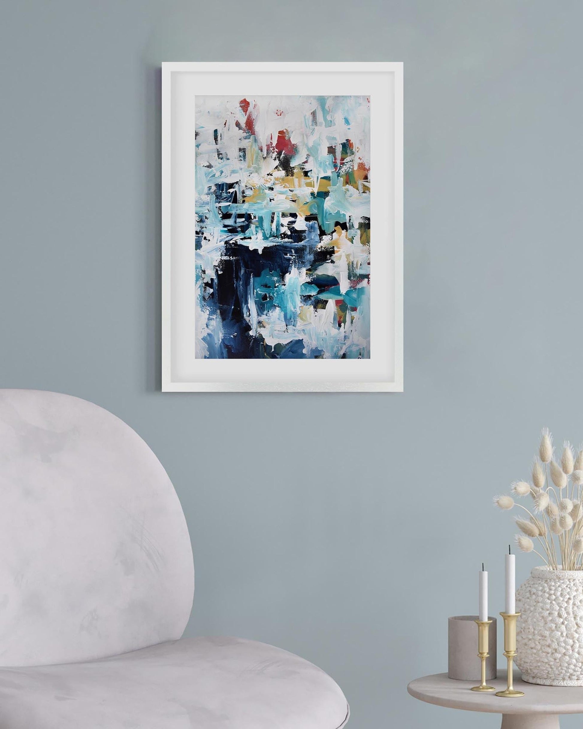 Framed abstract art piece on a light blue wall with a chair and small table in the foreground.