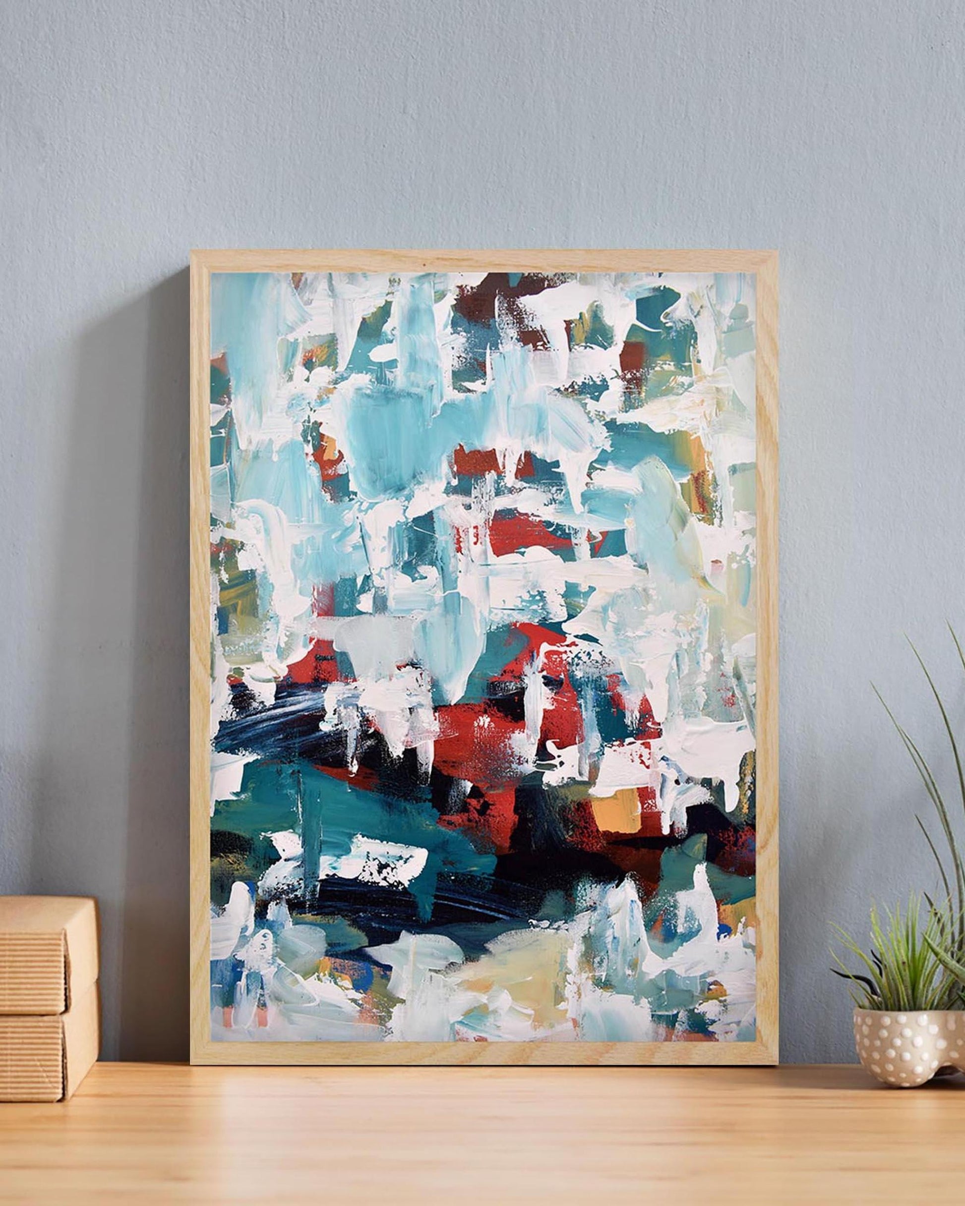 Abstract painting in a wooden frame on a wall with a plant and wooden side table.