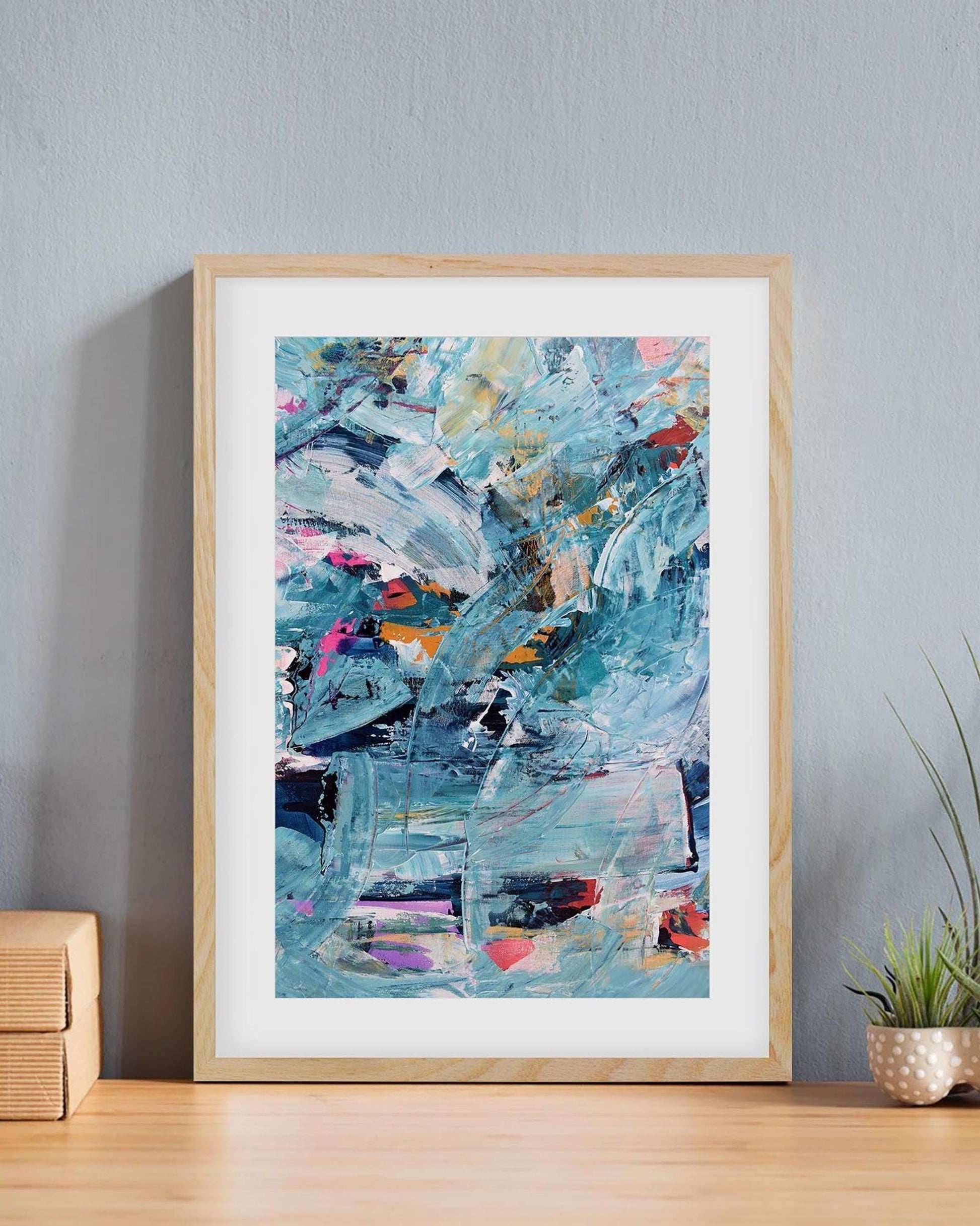 Framed abstract art print on a wall with a plant and wooden boxes below.