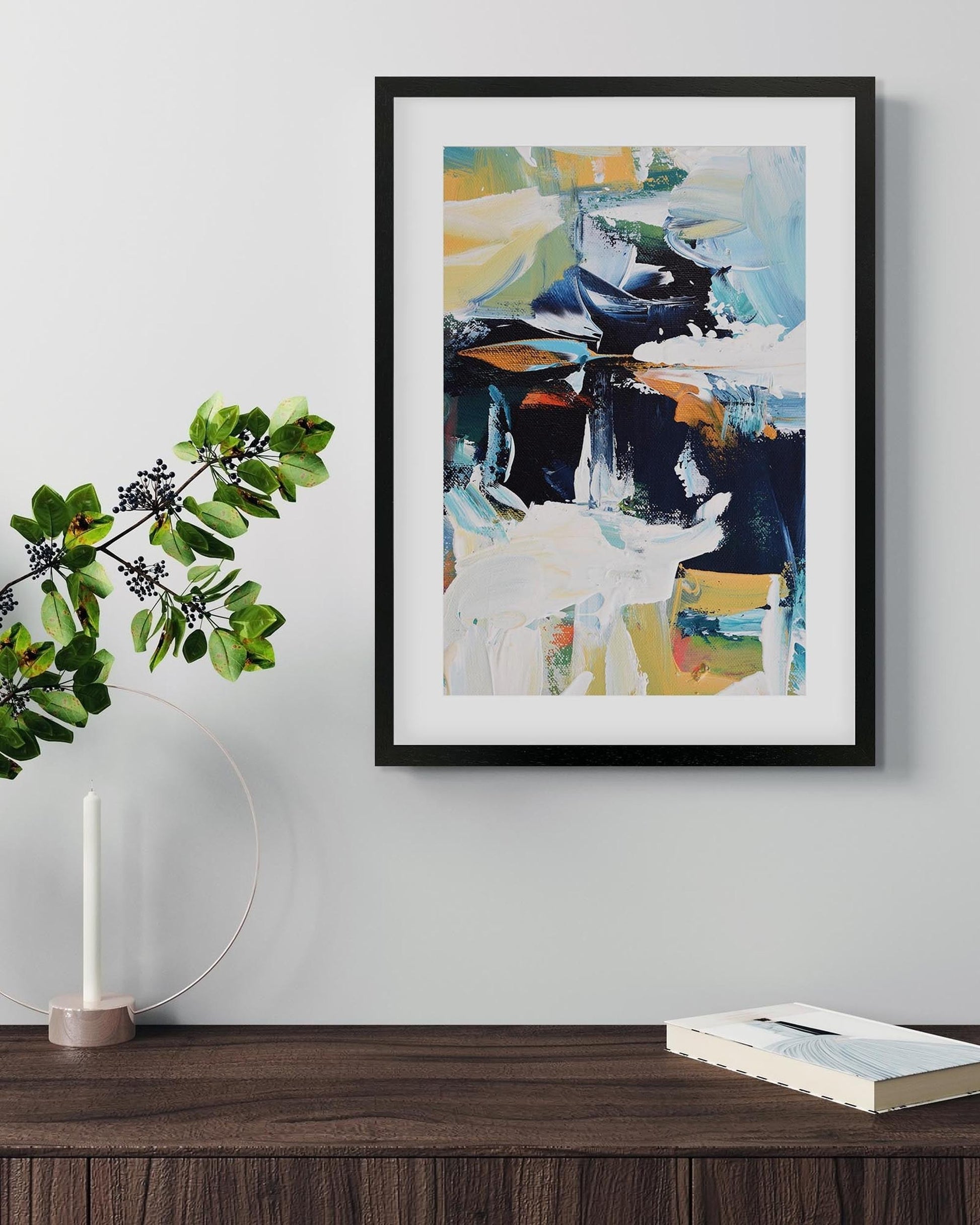 Framed abstract painting on a wall above a wooden surface with a candle and book.