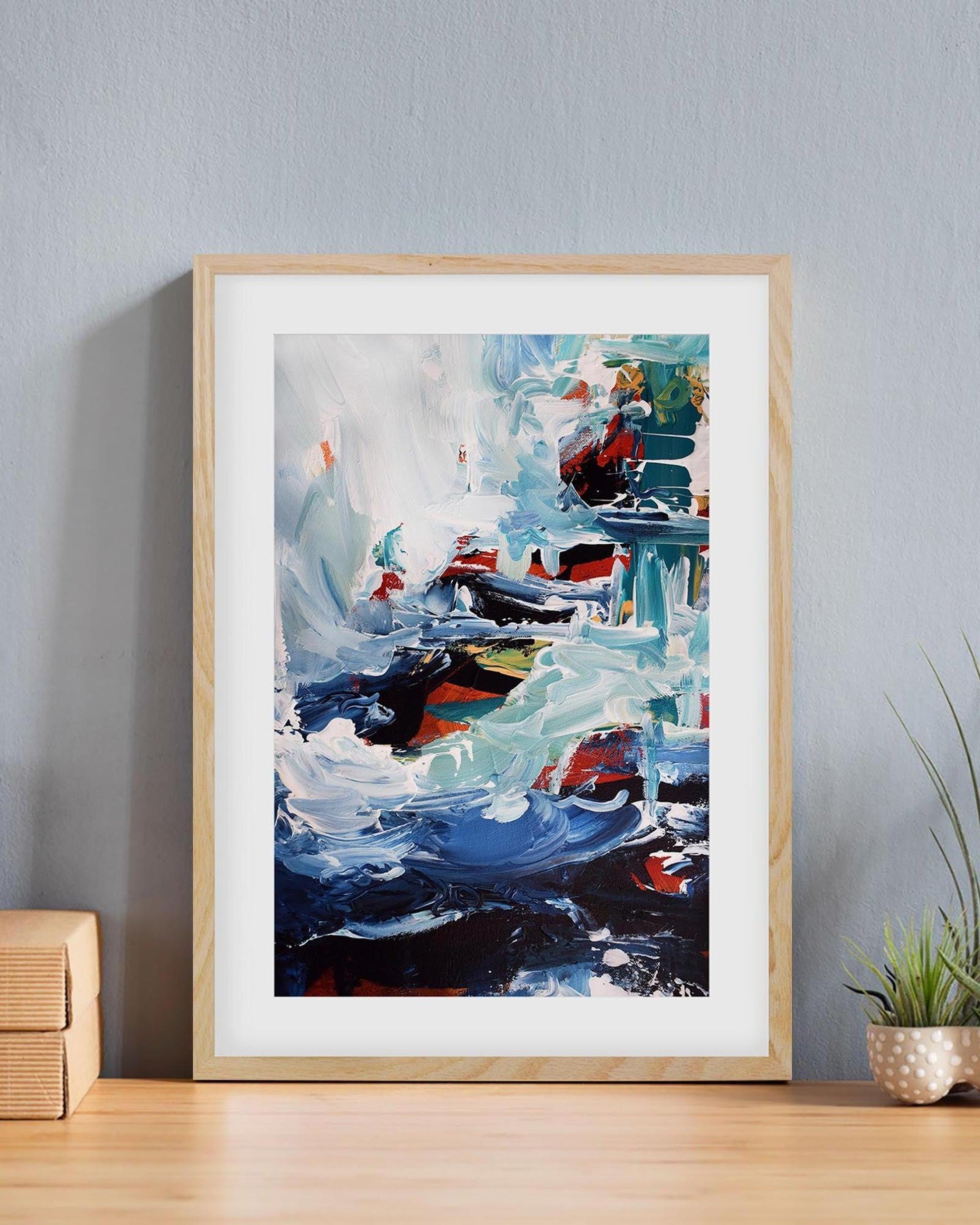 Framed abstract painting on a wall with a plant and wooden box in the foreground.