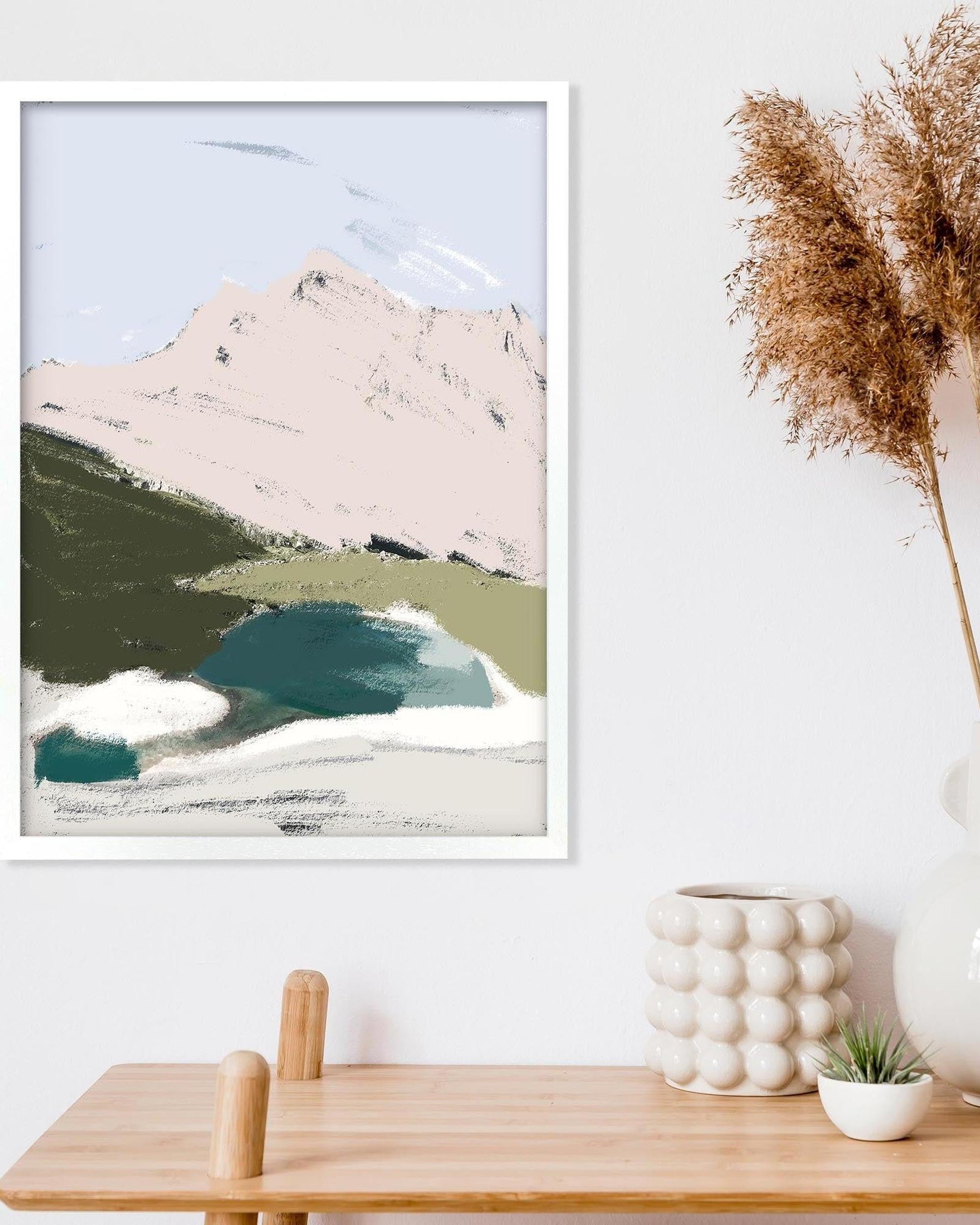 Framed abstract art print of a mountain landscape on a wall above a wooden table.