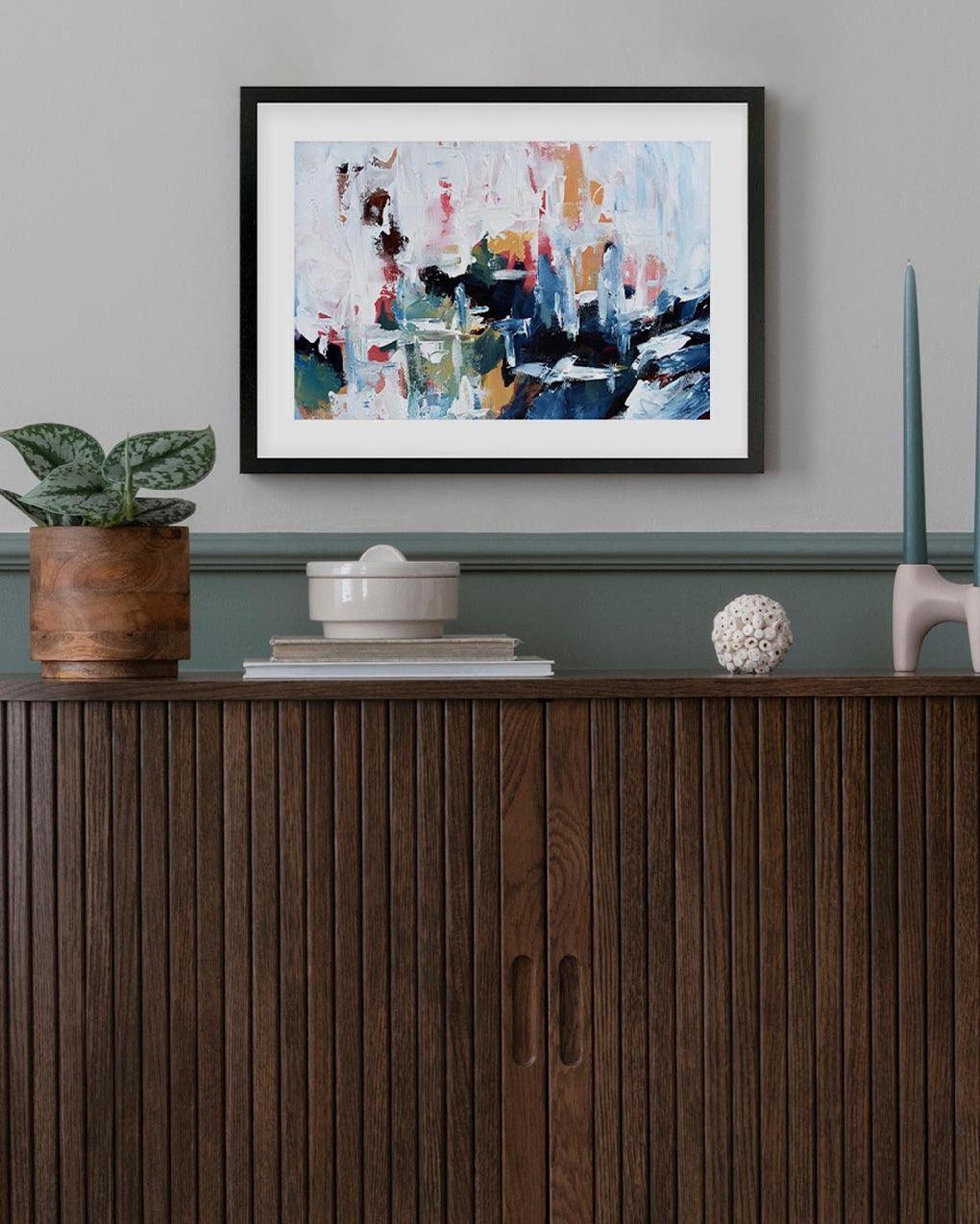 Framed abstract painting on a wall above a wooden cabinet with decorative items.