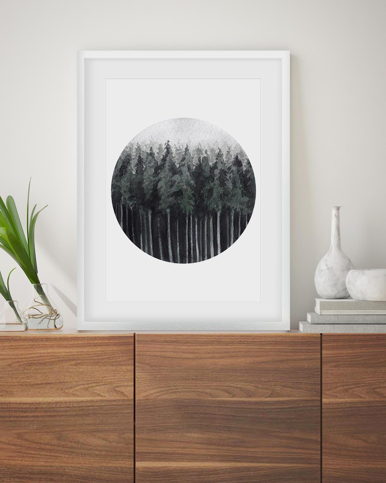 Framed circular artwork of a forest on a wall above a wooden cabinet.
