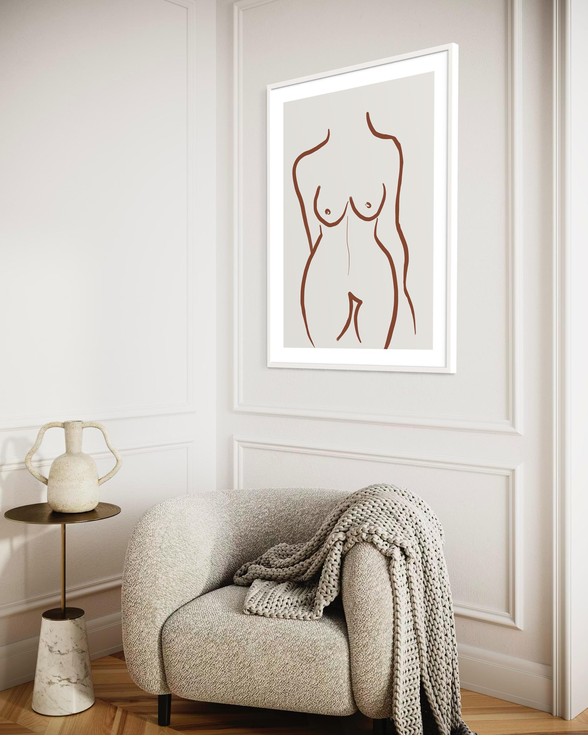 Framed abstract art of a nude figure on a wall above a cozy armchair with a blanket.