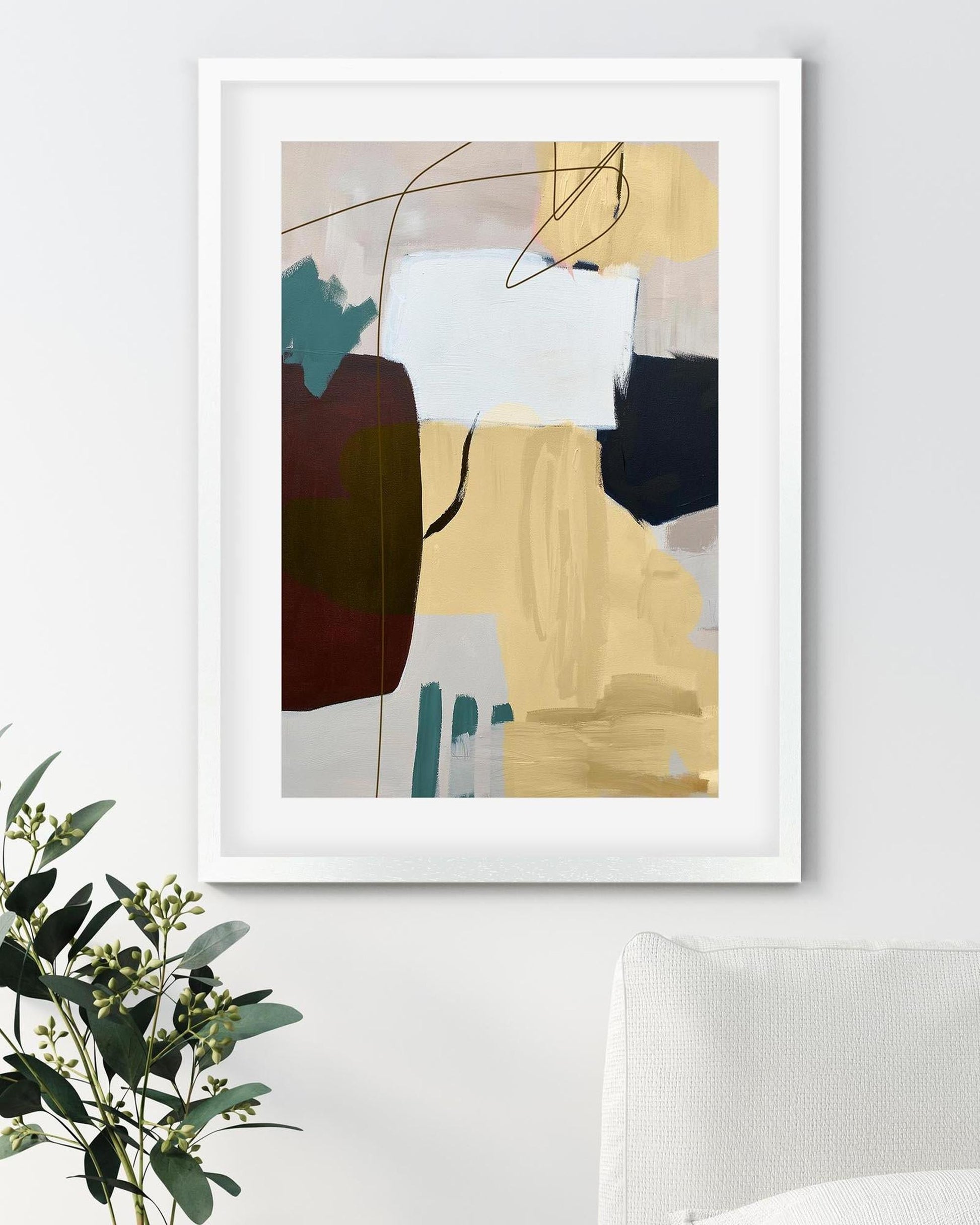 Abstract art print in a white frame on a wall with a plant and chair in the foreground.