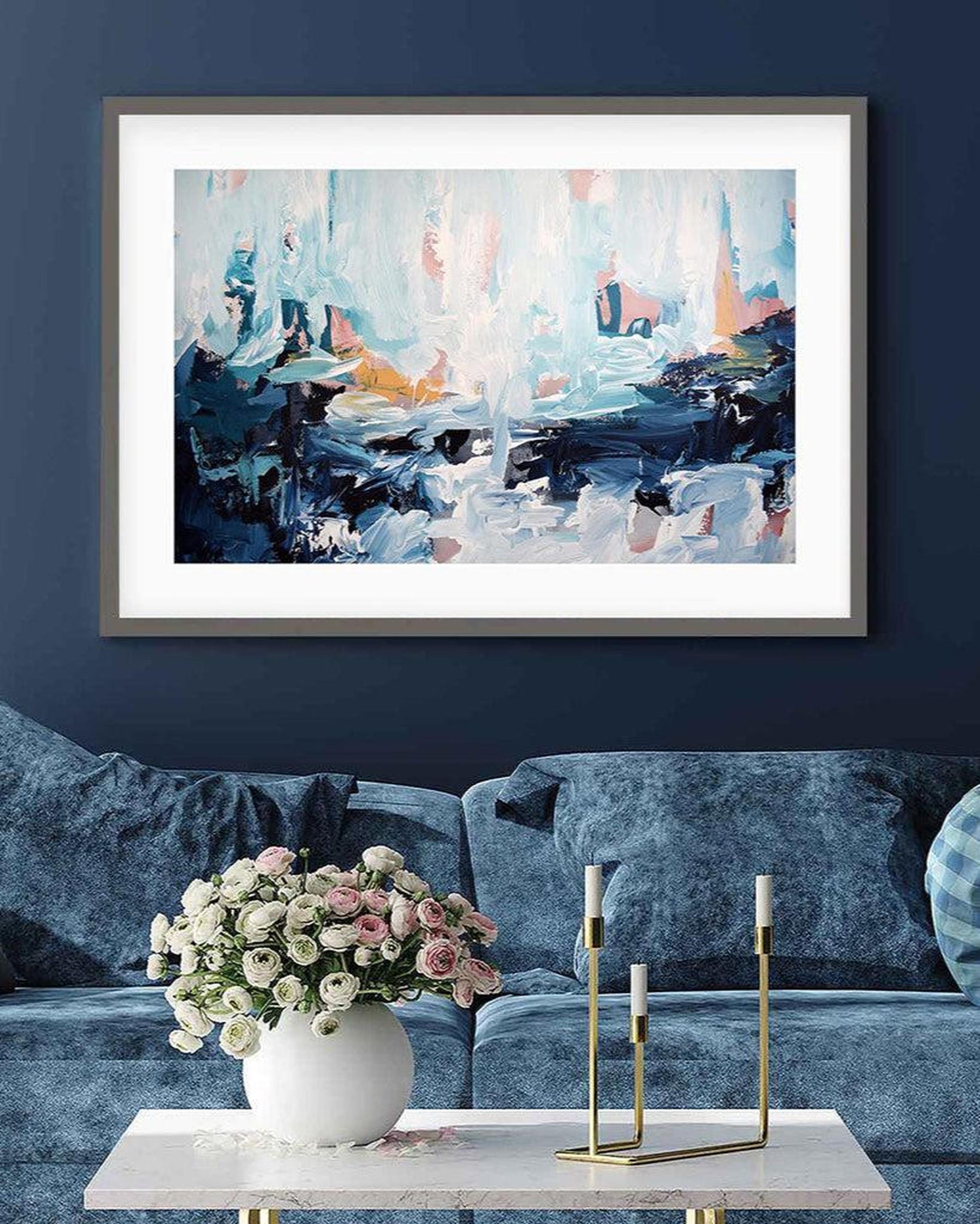 Framed abstract painting on a blue wall above a sofa with a vase of flowers and candlesticks.