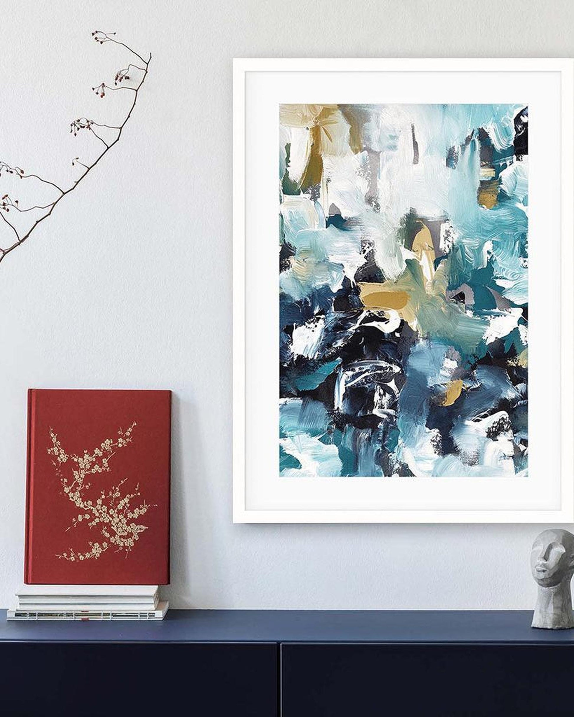 Framed abstract art piece on a wall with a red book and sculpture below.