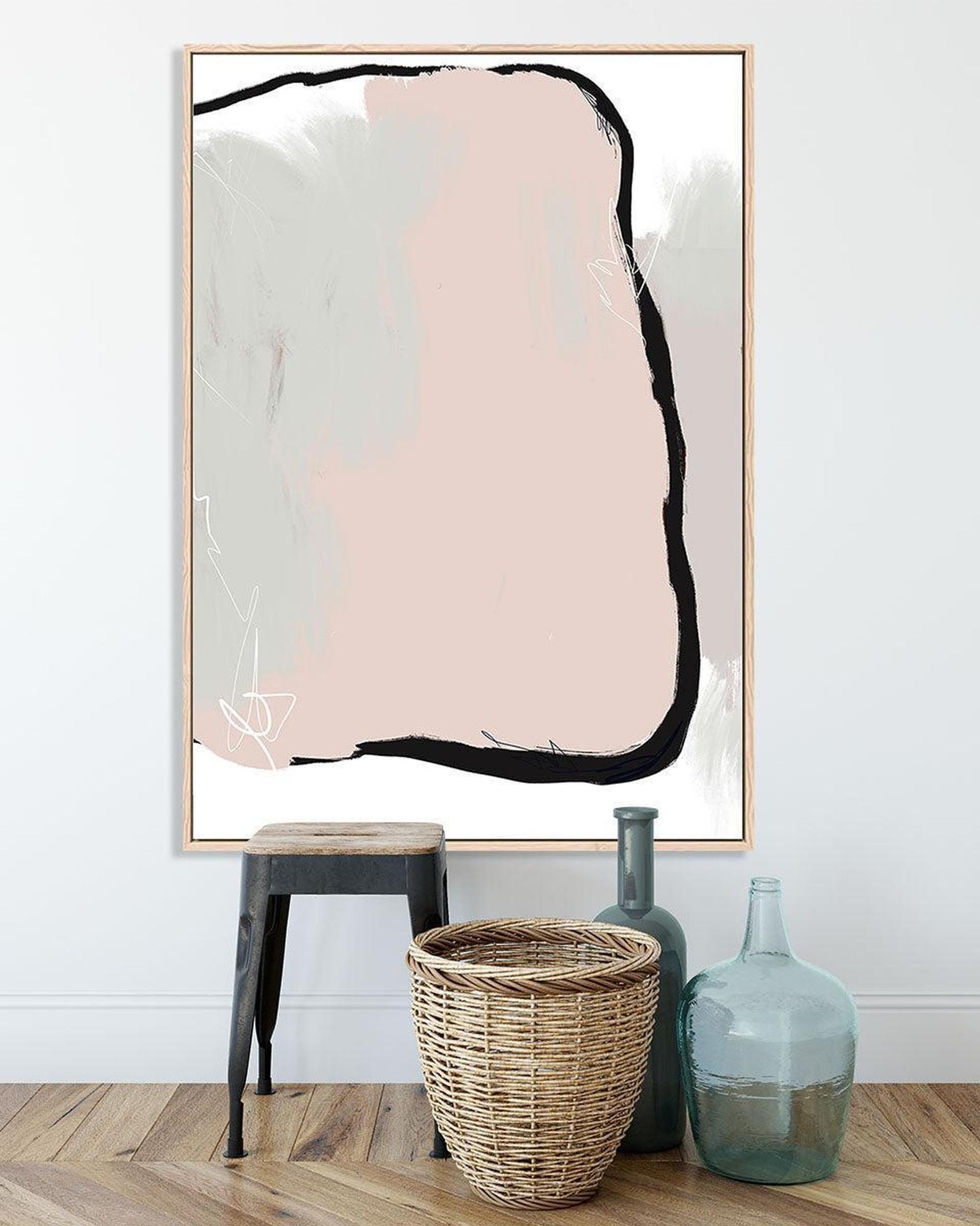 Abstract art print with a pink and black shape in a gold frame, placed on a wooden floor with decorative items.