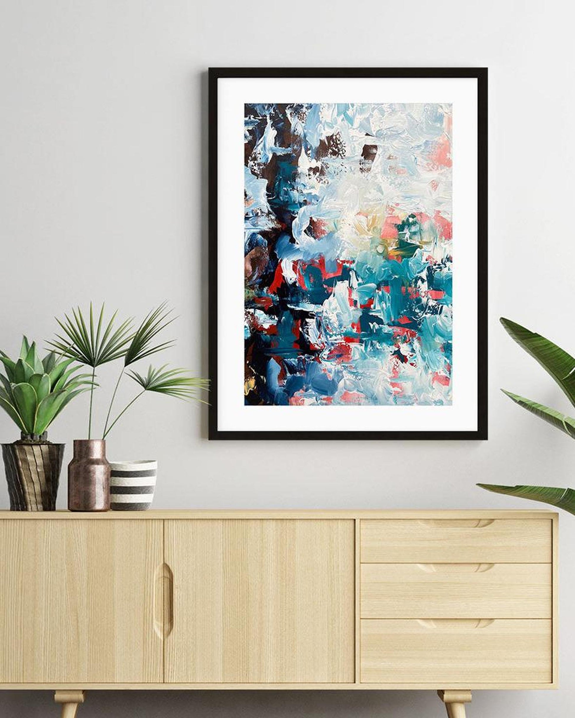 Framed abstract painting on a wall above a wooden cabinet with plants.