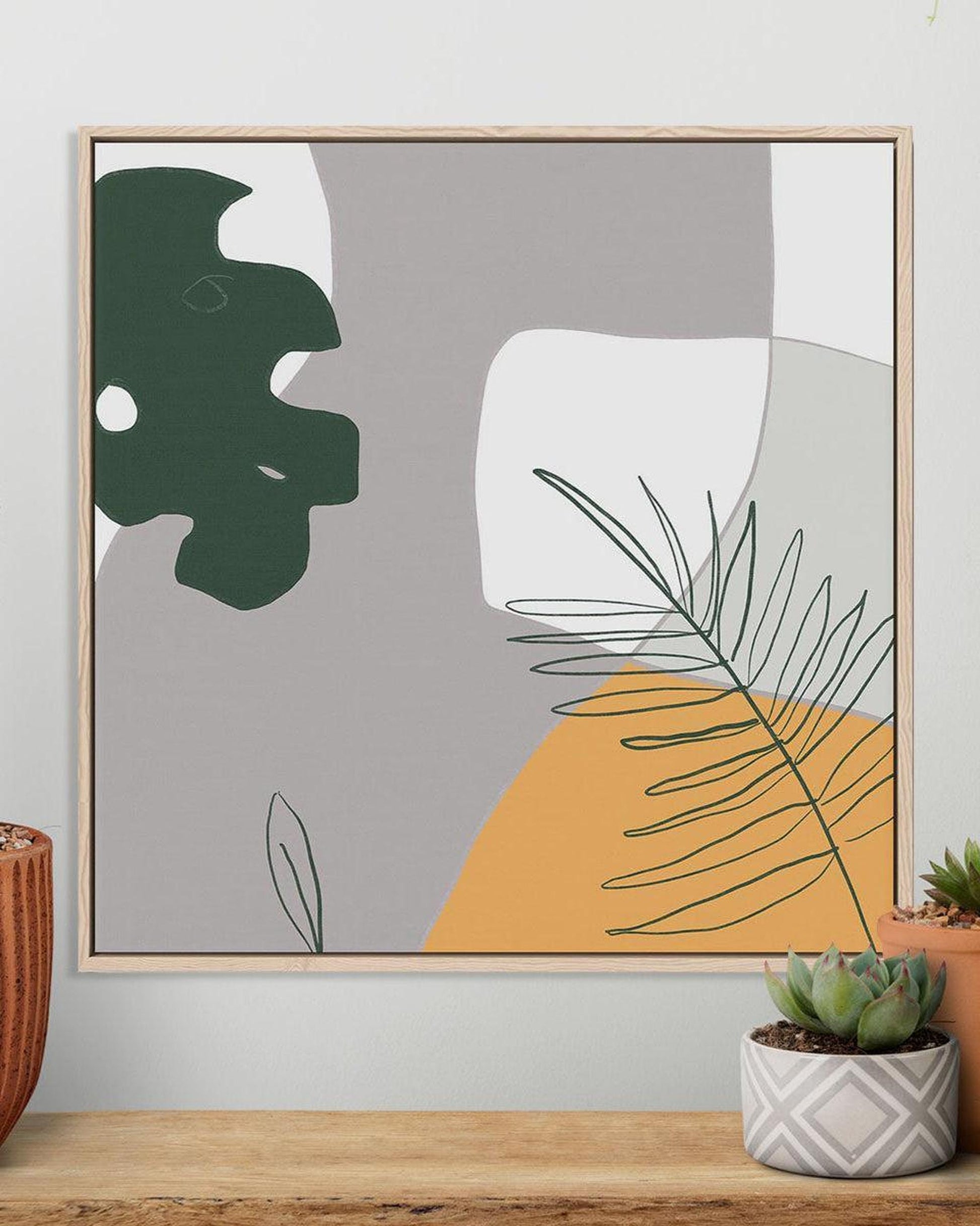 Framed abstract art with green and yellow shapes on a gray background, displayed on a wall above a wooden shelf.