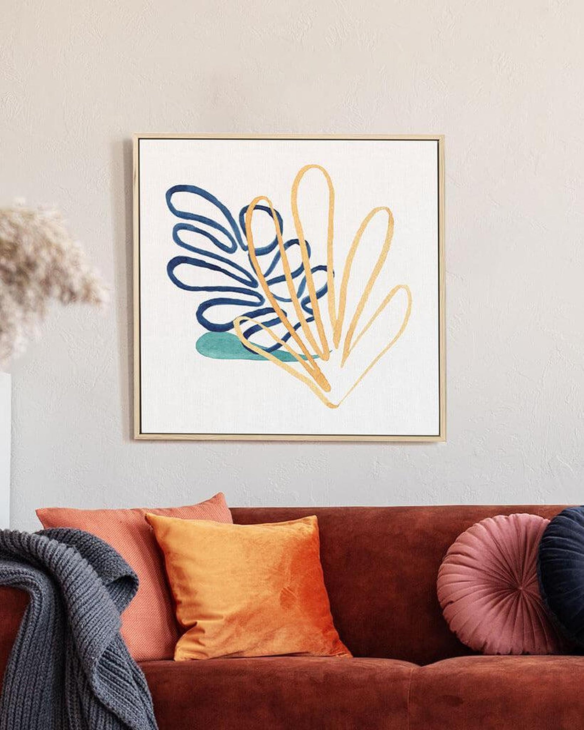 A framed canvas print featuring abstract botanical art in a vibrant color palette, displayed above a brown sofa with orange and pink cushions.