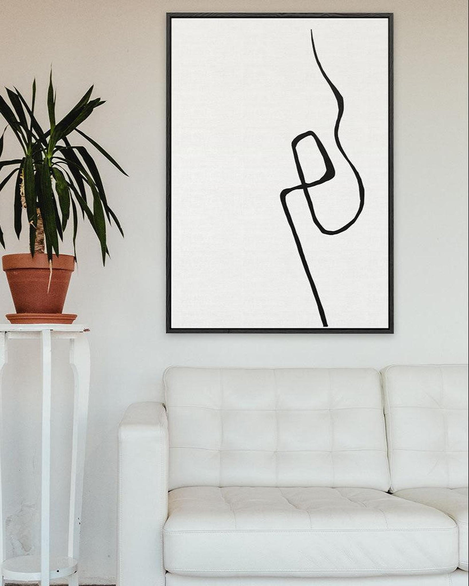 Abstract line art in a black frame on a white wall above a white sofa with a plant beside it.