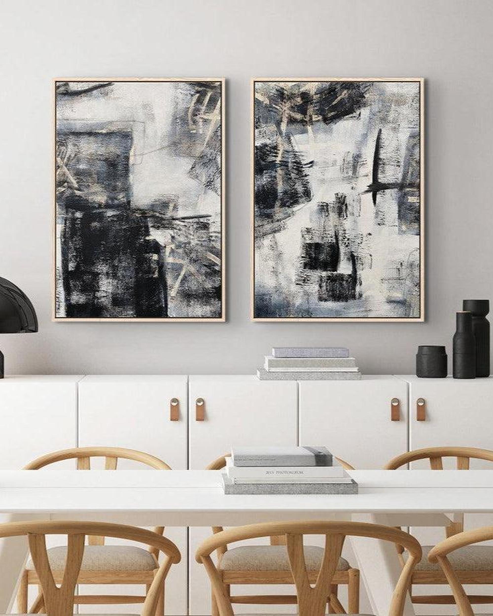 A set of two contemporary abstract art canvases with grey tones and light gold undertones, displayed above a white console table in a room with wooden chairs and a window.