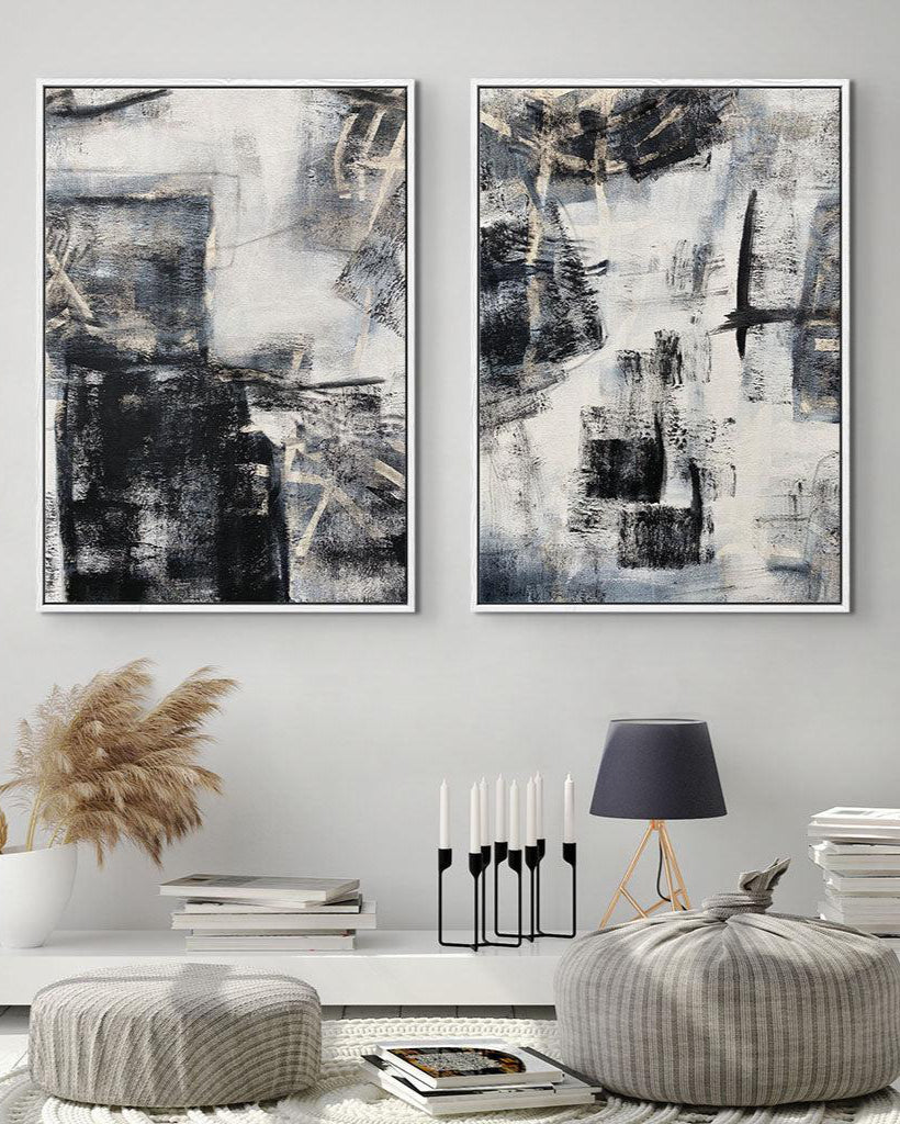 A set of two contemporary abstract art canvases with grey tones and light gold undertones, displayed above a white console table in a room with wooden chairs and a window.