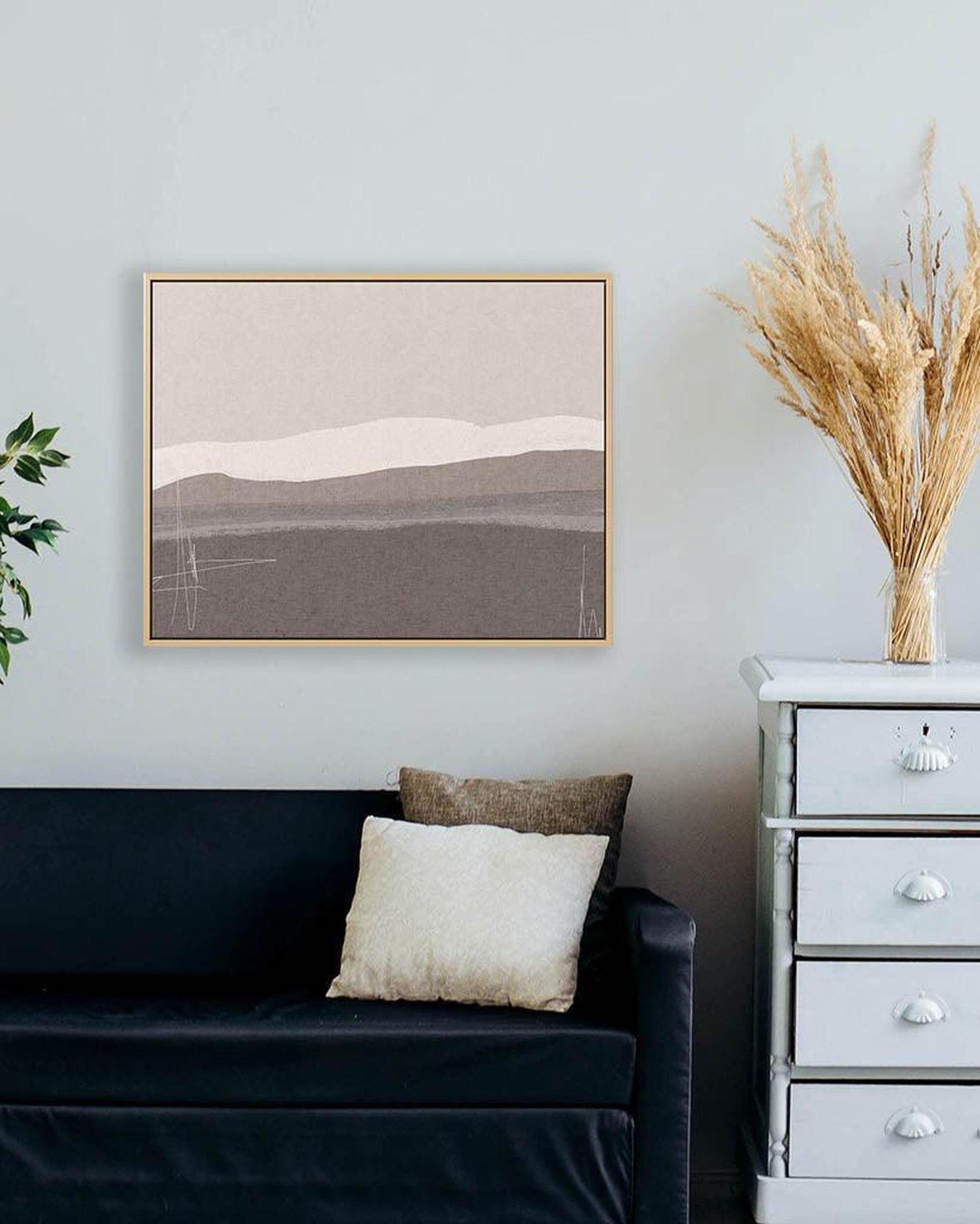 Living room with a dark blue sofa, white pillow, framed landscape art, and a white dresser with dried plants.
