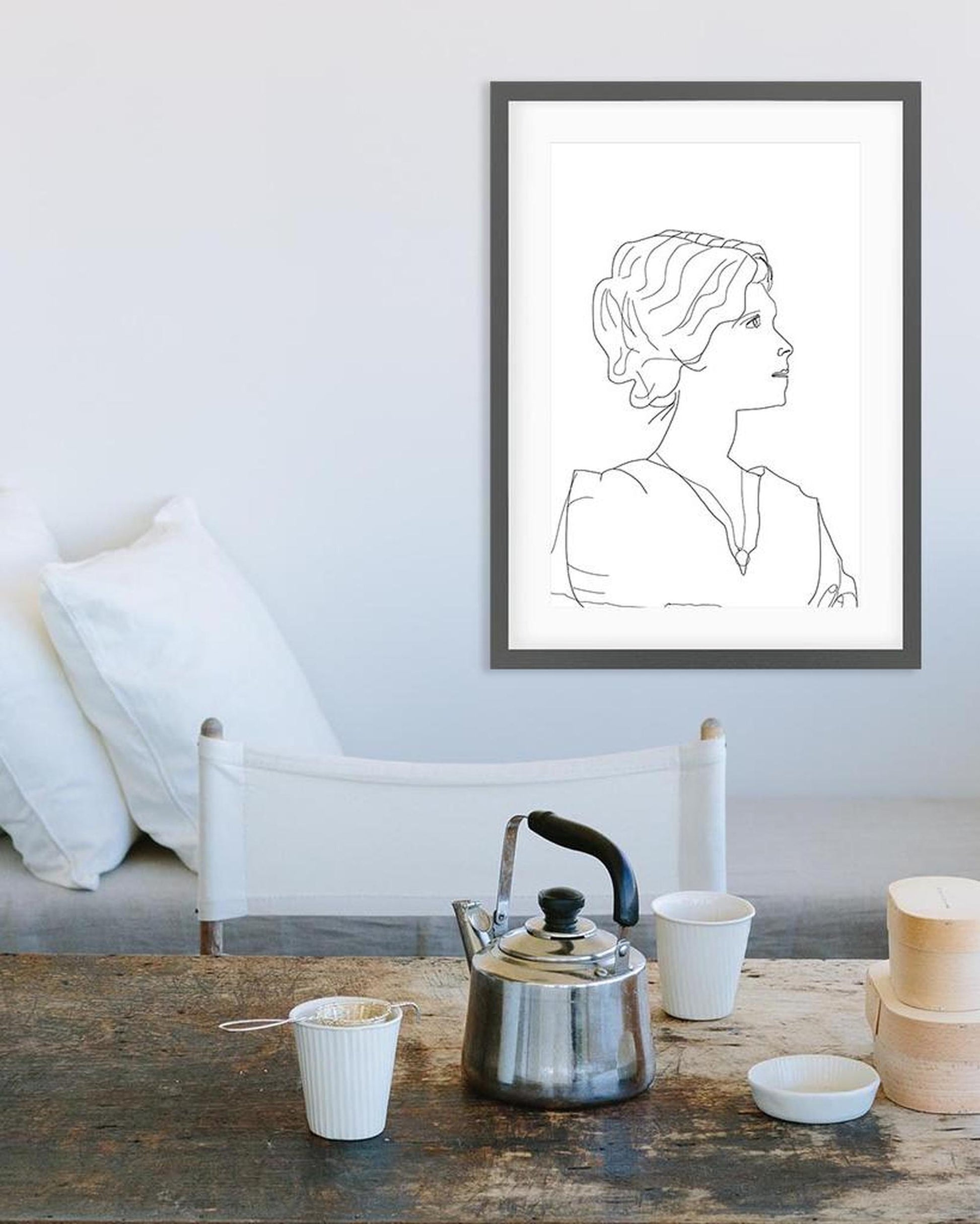 Framed line art of a woman's portrait above a rustic wooden table with tea-making items.